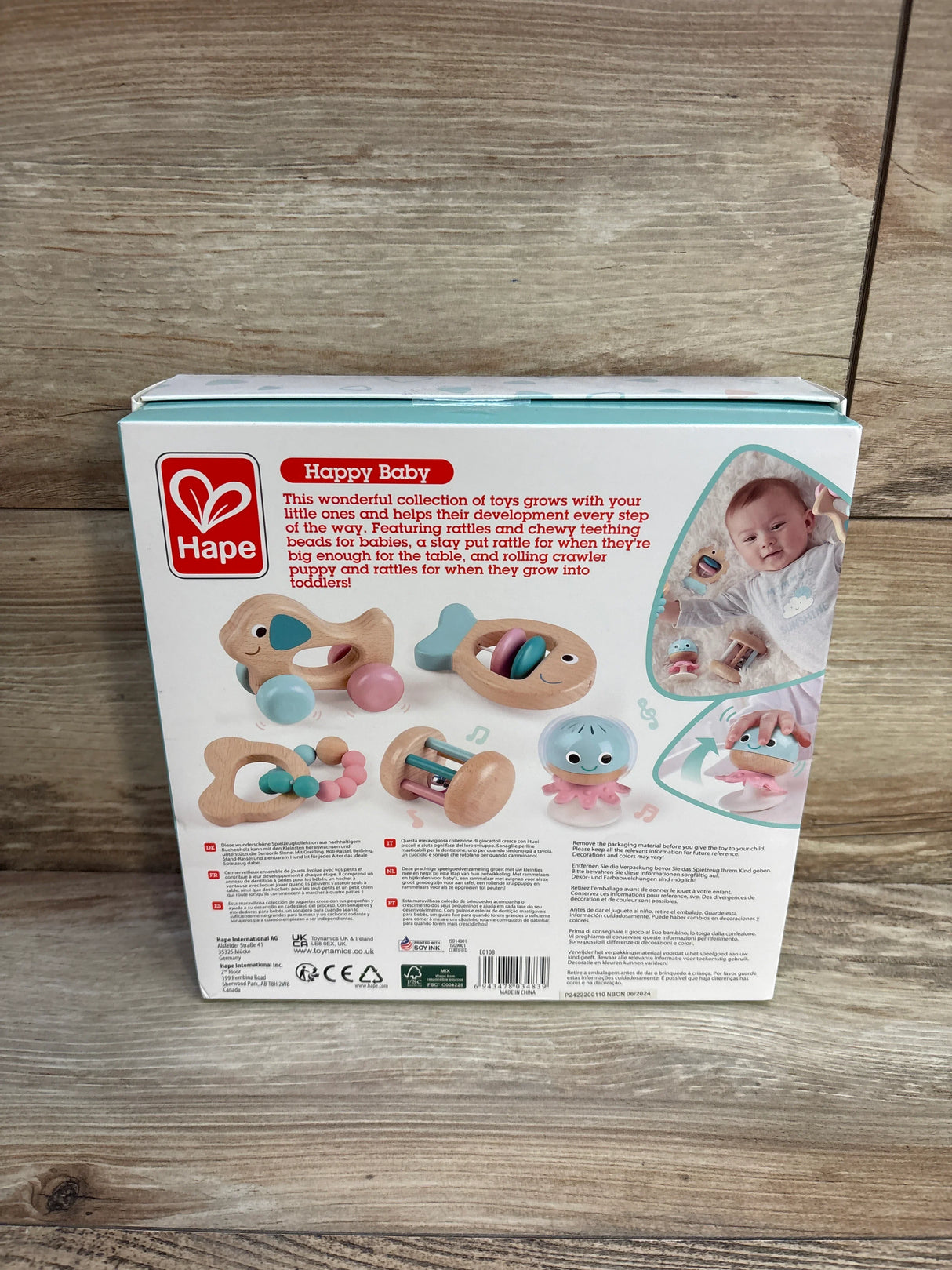 Hape Happy Baby toy set box showing wooden baby rattles, teething toys, and infant photo on wood background