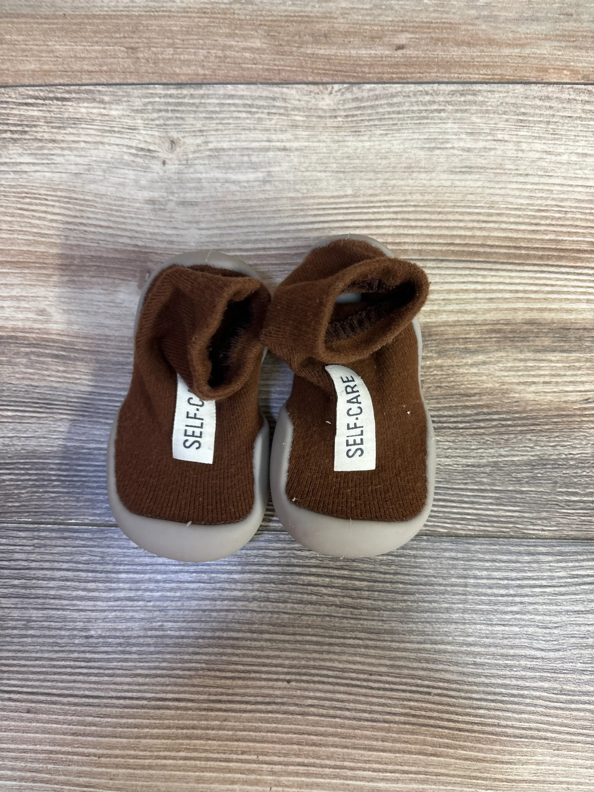 Top view of brown Tiny Explorings self-care sock shoes showcasing gray soles and SELF-CARE labels on a wood-grain background