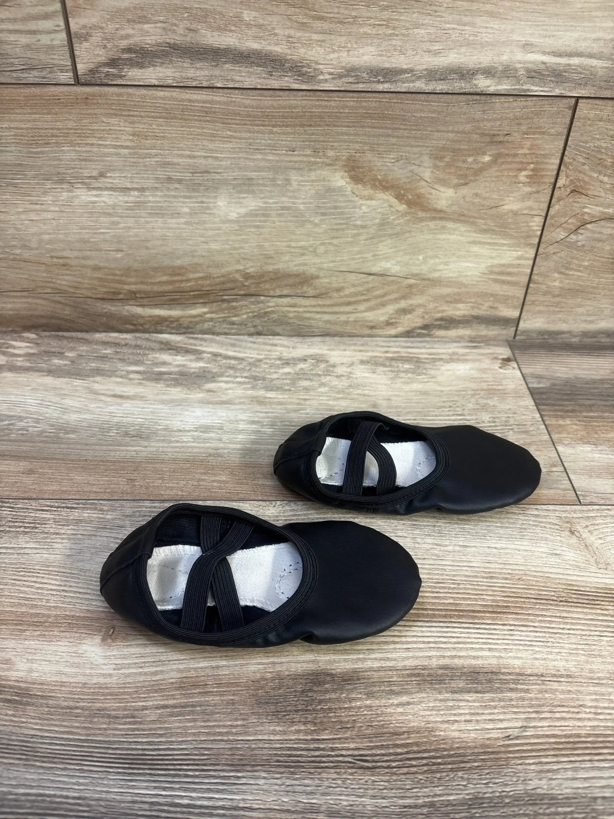 Top-down view of black Stelle toddler ballet shoes showing crossed elastic straps and white canvas lining on wood floor background