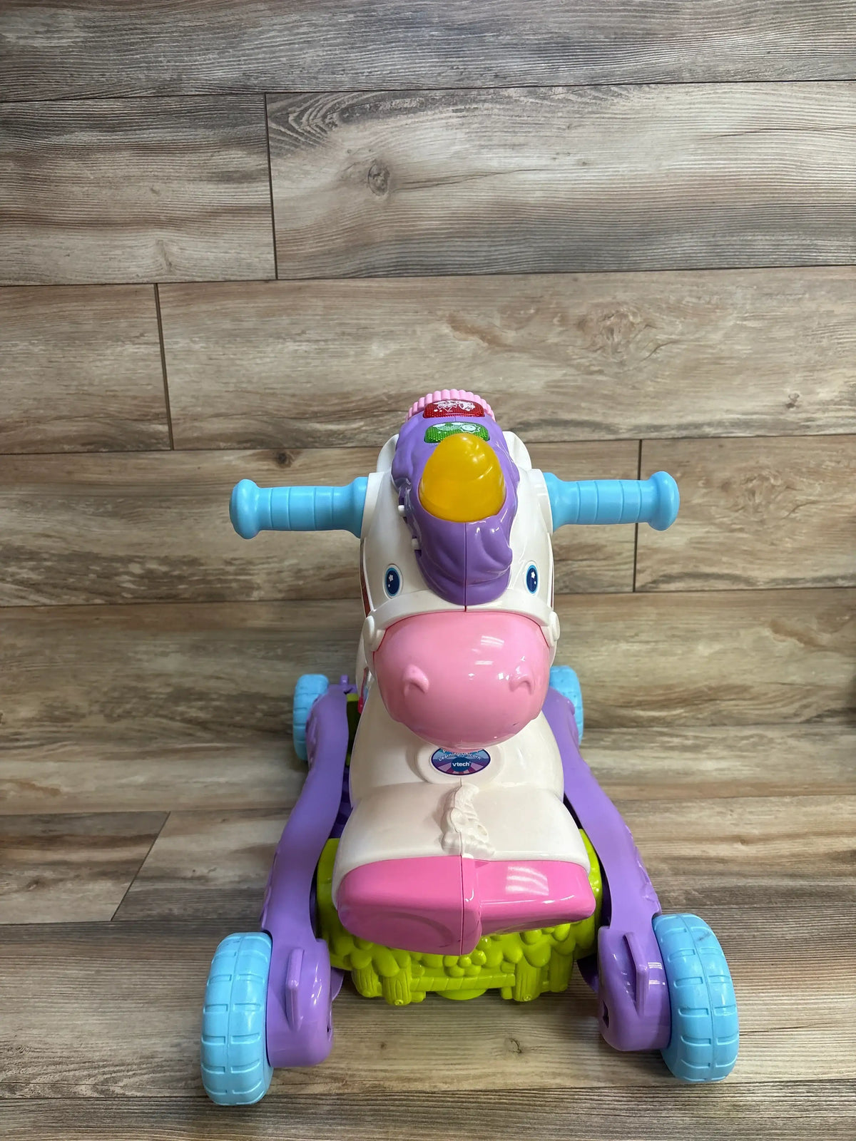 Front view of VTech Prance and Rock Learning Unicorn showcasing white purple and pink ride-on with blue handles and rocker base on wood plank floor