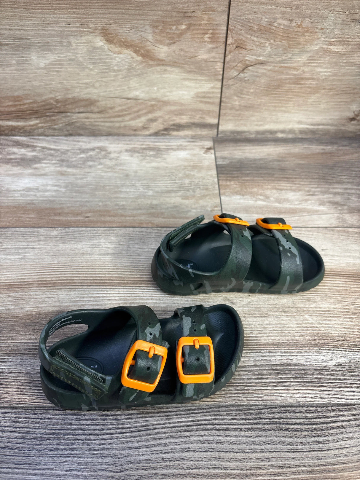 Angled top view of toddler camo Cat & Jack footbed sandals showing bright orange buckles on a wood-plank background, size 8C