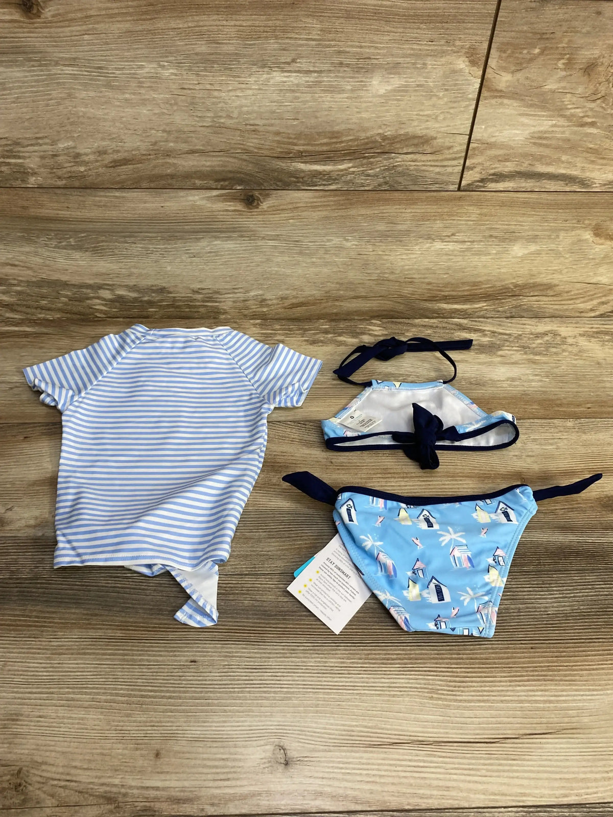 Flat lay of toddler blue halter bikini and matching rash top showing beach print and navy ties on wood background