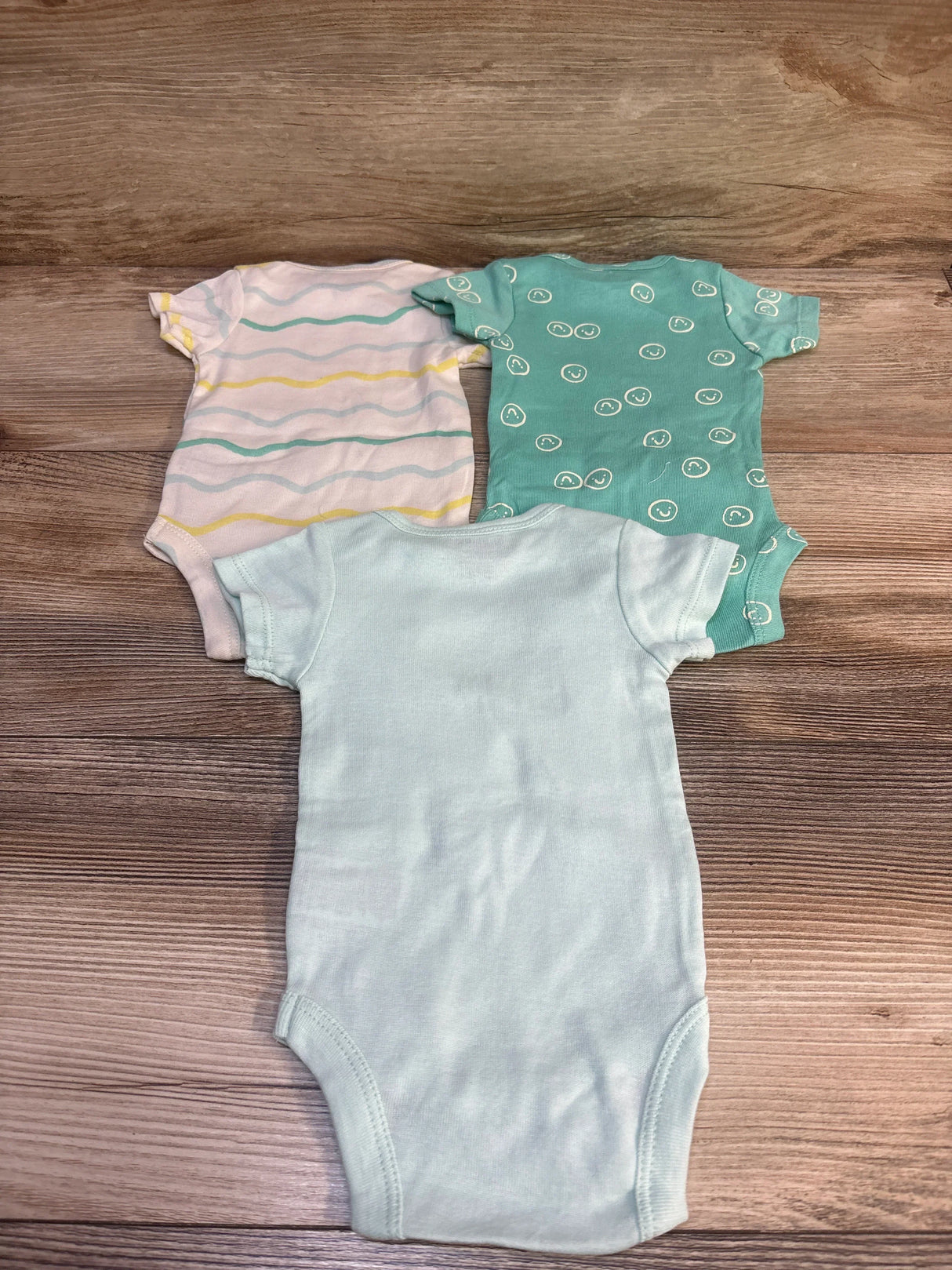 Top view of three newborn Onesies Brand bodysuits—solid aqua, aqua smiley print, and white with aqua/yellow stripes on wood