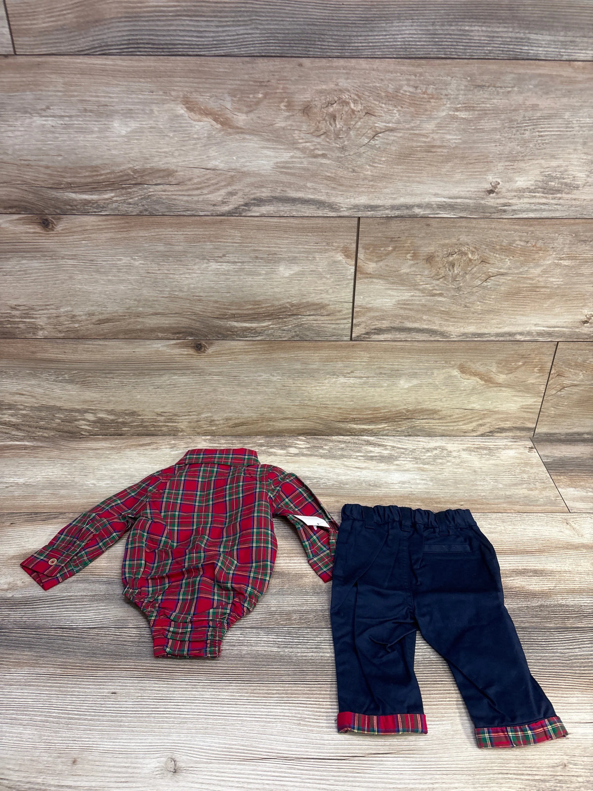 Top down view of a red plaid two piece baby outfit set with long sleeve bodysuit and navy pants with plaid cuffs on wood
