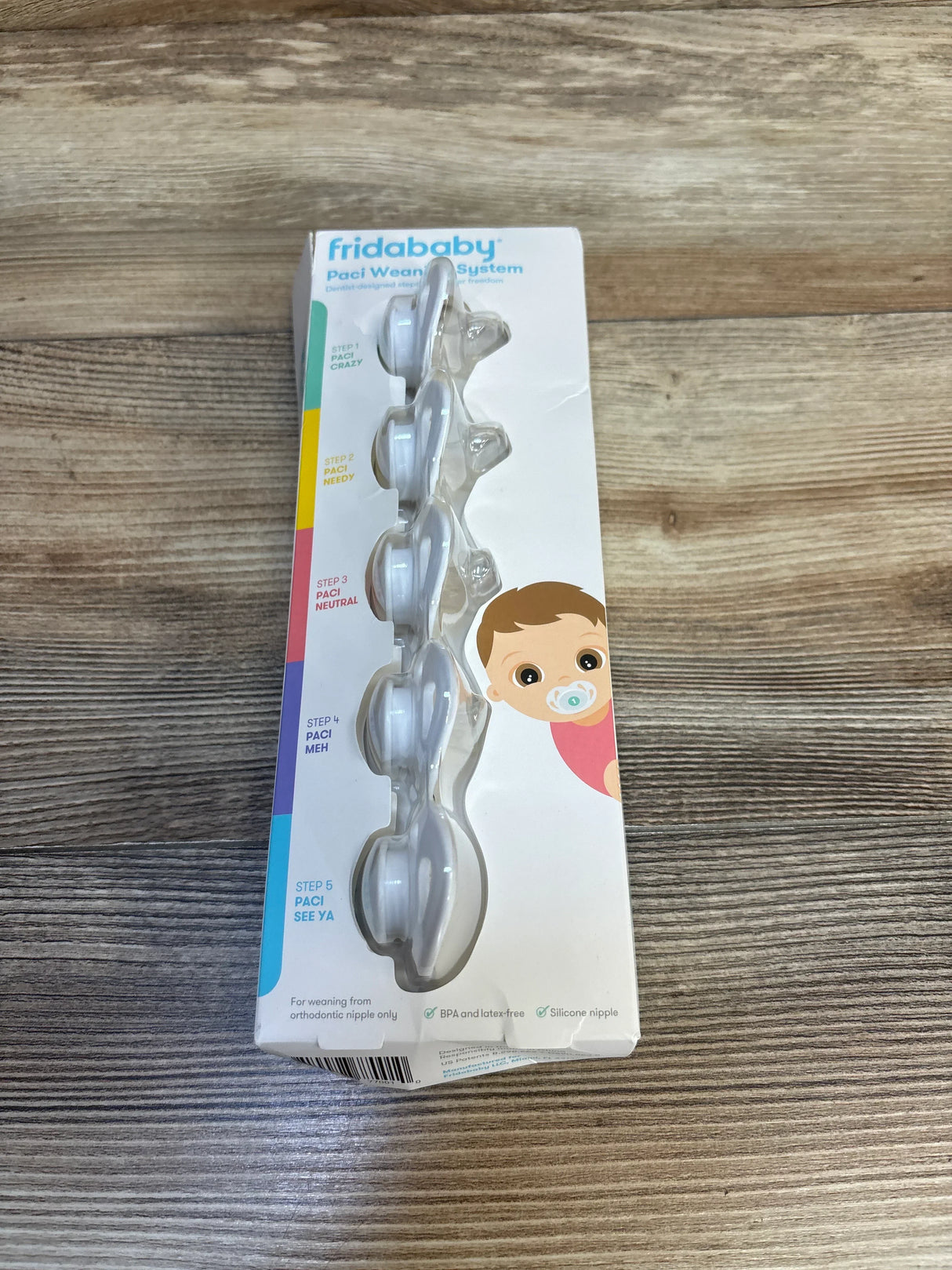 Front view of NEW Frida Baby Paci Weaning System 5ct showcasing five-stage clear pacifier set in sealed retail box on wood plank floor