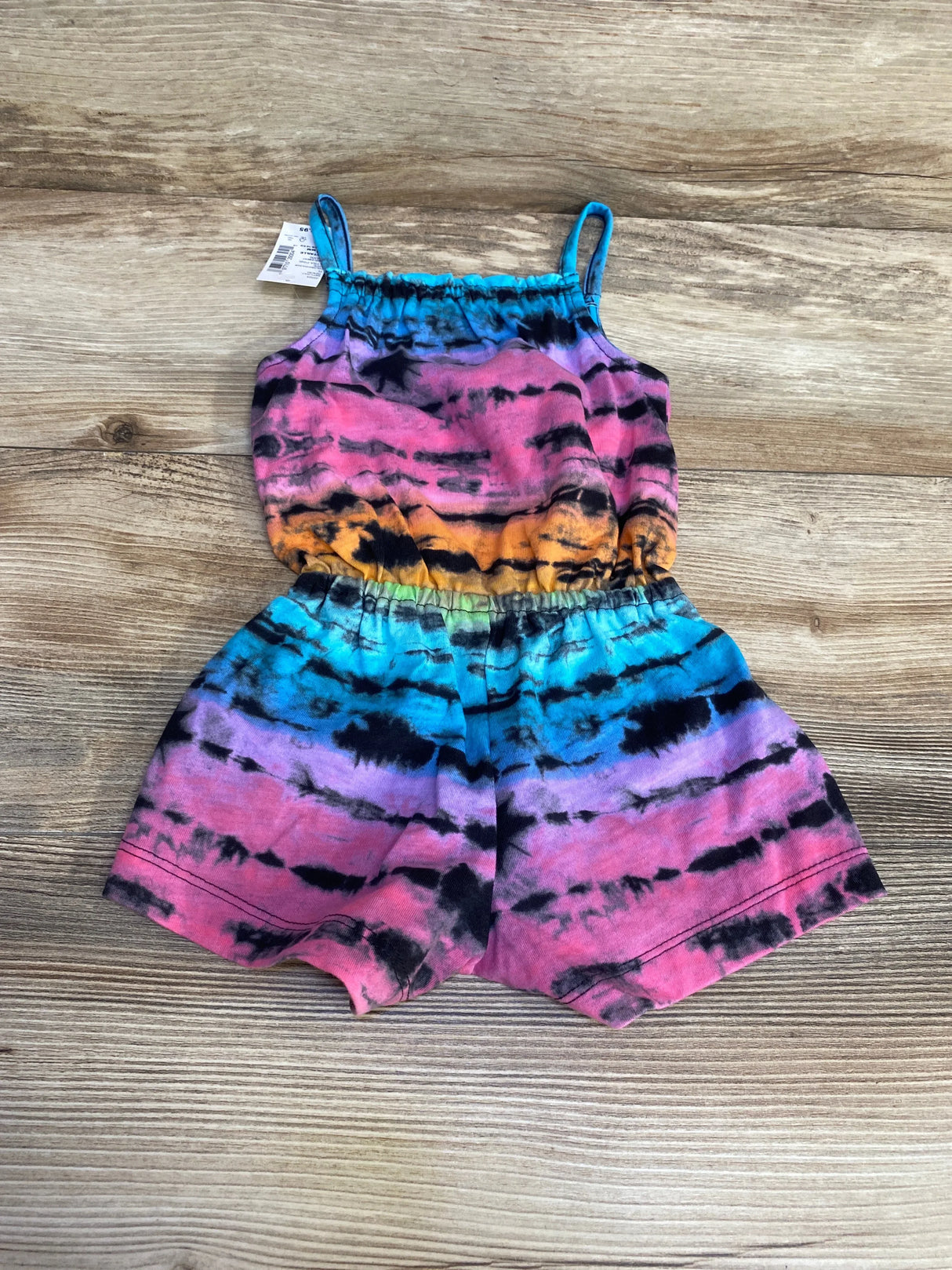 Flat-lay of Children's Place tie-dye romper showing thin shoulder straps, elastic waist, and multicolor stripes on wood background