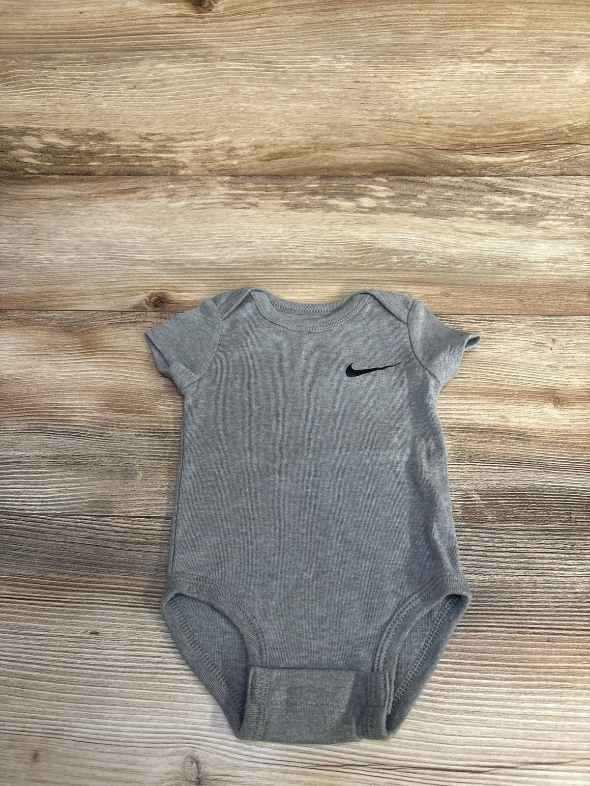 Top down view of grey Nike baby bodysuit size 6m showing short sleeves and small black Swoosh logo on chest on wood grain background