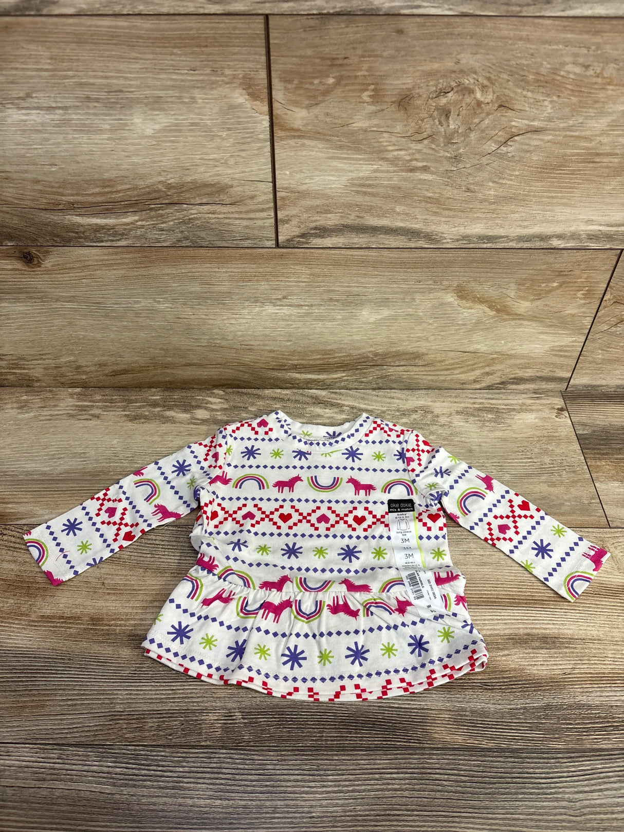 Front view of Okie Dokie unicorn fair isle peplum shirt in white, size 3M, with long sleeves on a wood floor