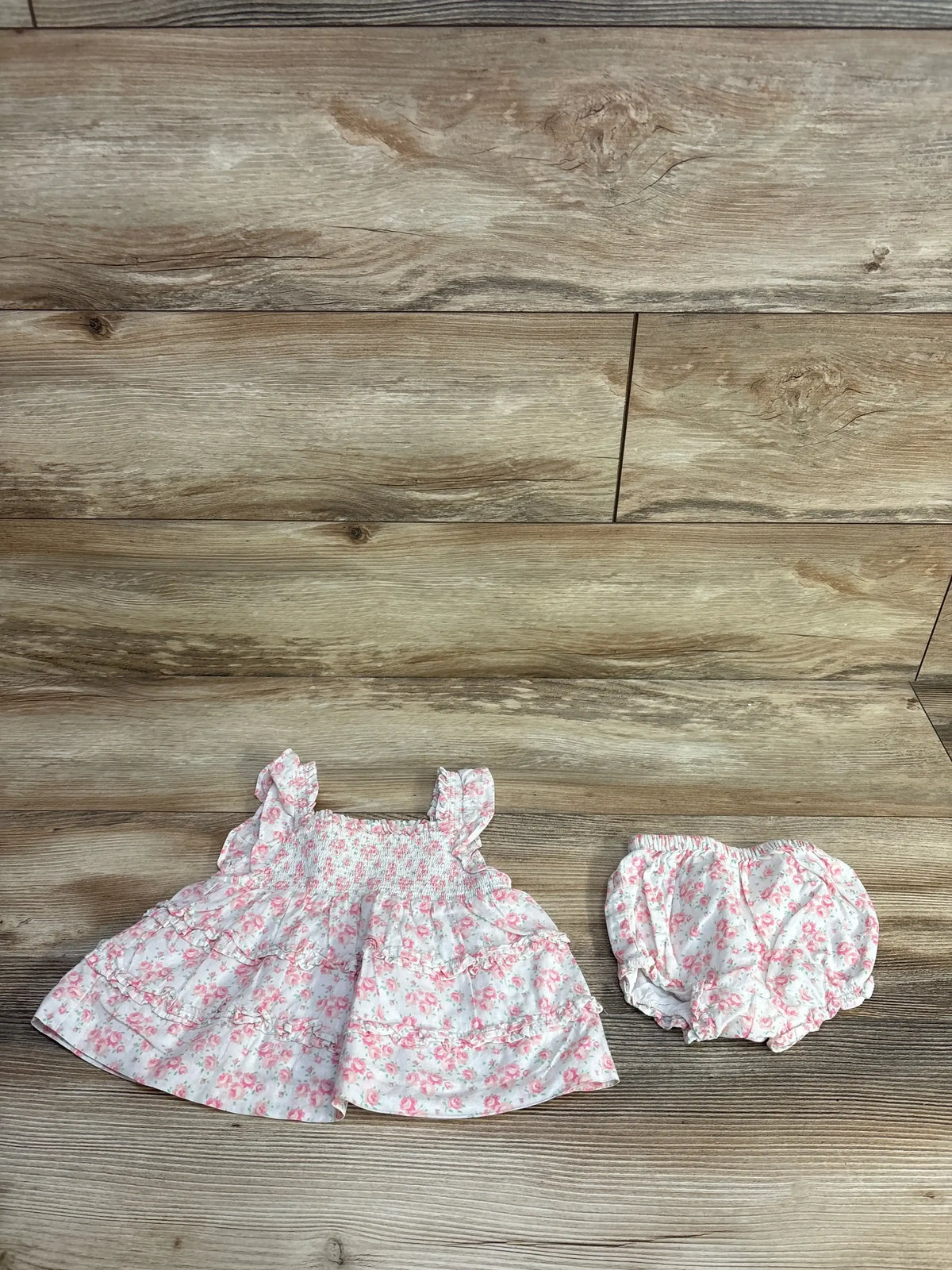 Flat lay of Janie & Jack 2pc The Emily Floral Smocked Set sz 6-12m white dress with pink floral print and matching bloomers on wood floor