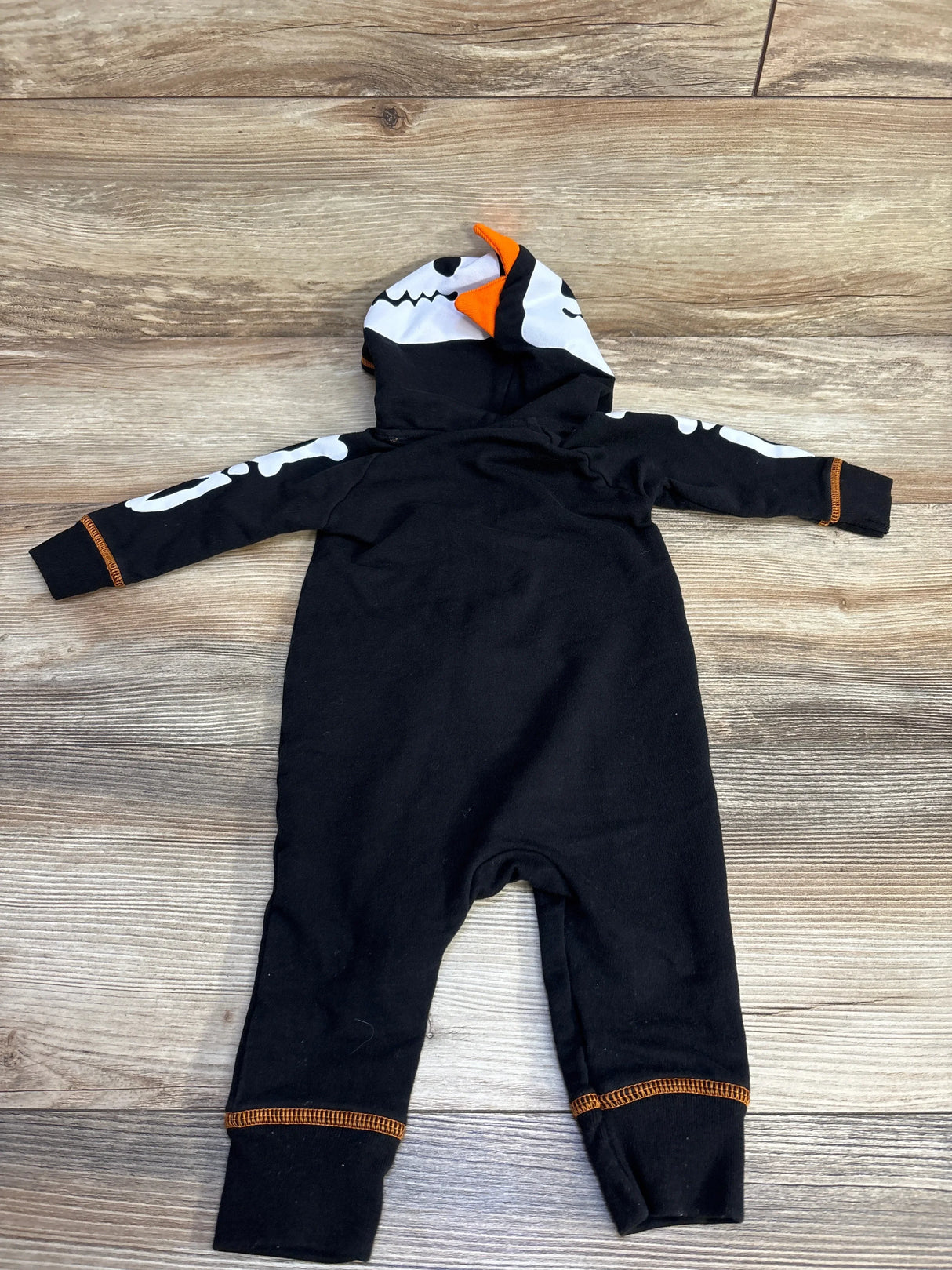 Black skeleton Halloween baby onesie with hood, orange trim, and bone print on sleeves, laid flat on wood floor.