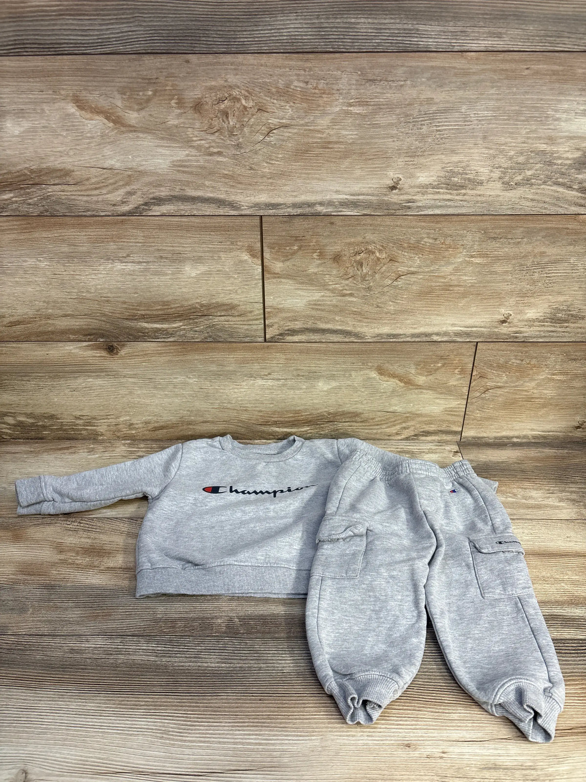 Top-down view of Champion 2-piece pullover sweatshirt and joggers set showcasing grey fabric, logo, and cargo pockets on wood flooring