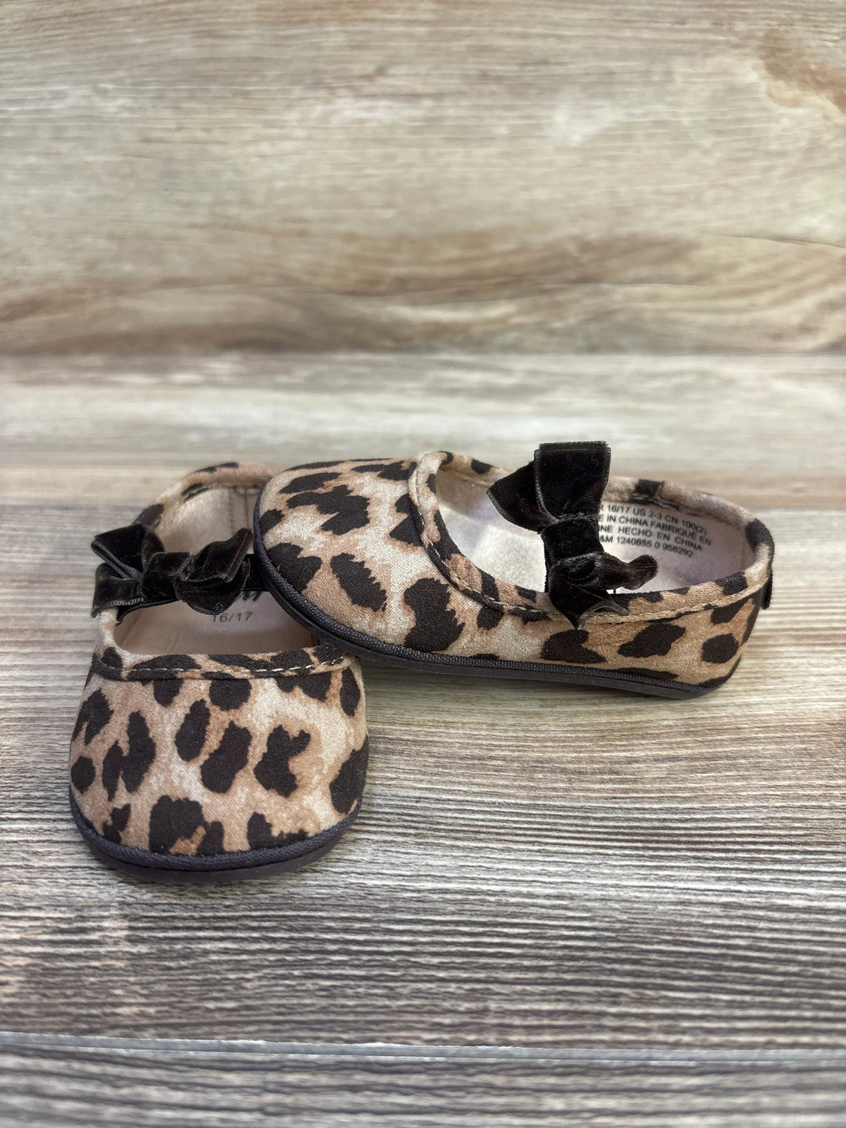 Front and side view of leopard-print baby ballet flats showcasing black velvet bows and rounded toes on a light wood surface
