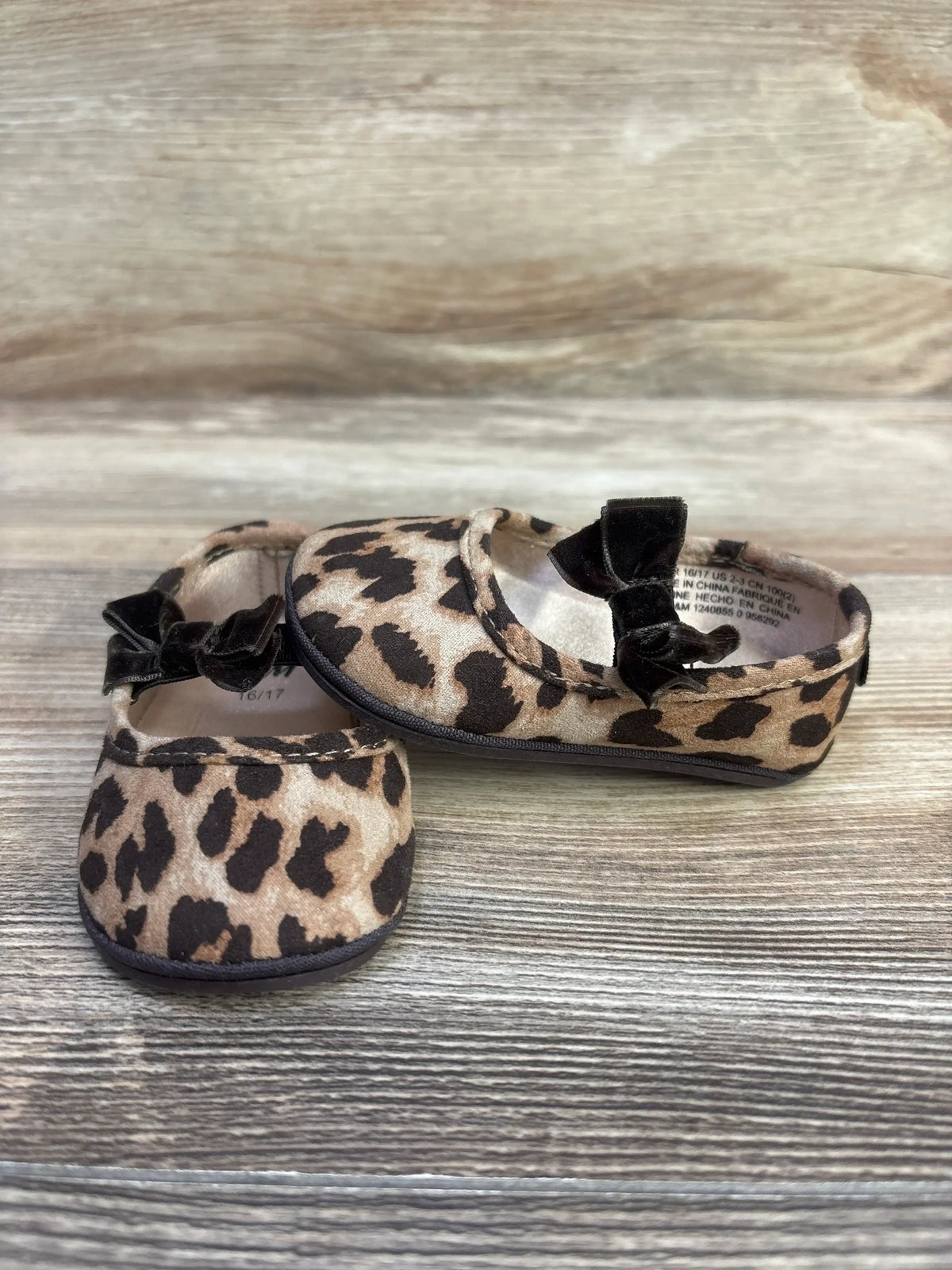 Front and side view of leopard-print baby ballet flats showcasing black velvet bows and rounded toes on a light wood surface