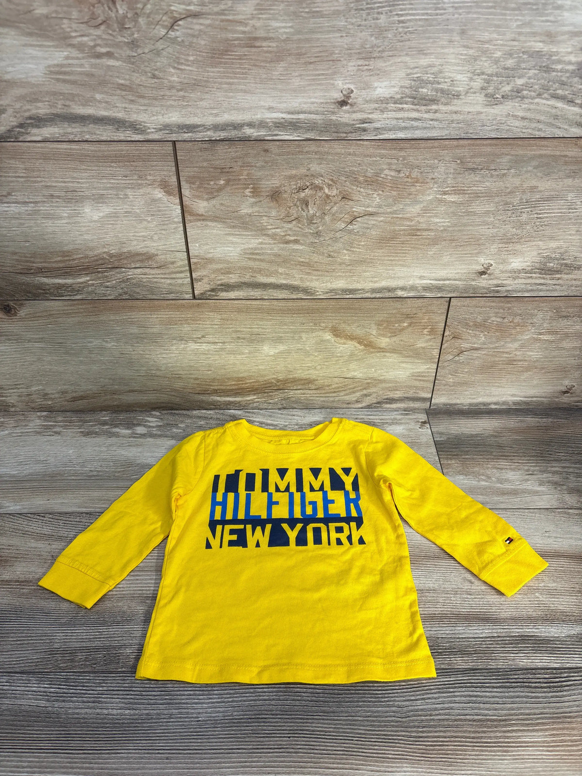 Front view of a bright yellow Tommy Hilfiger logo shirt showcasing long sleeves and New York chest lettering on a wood floor