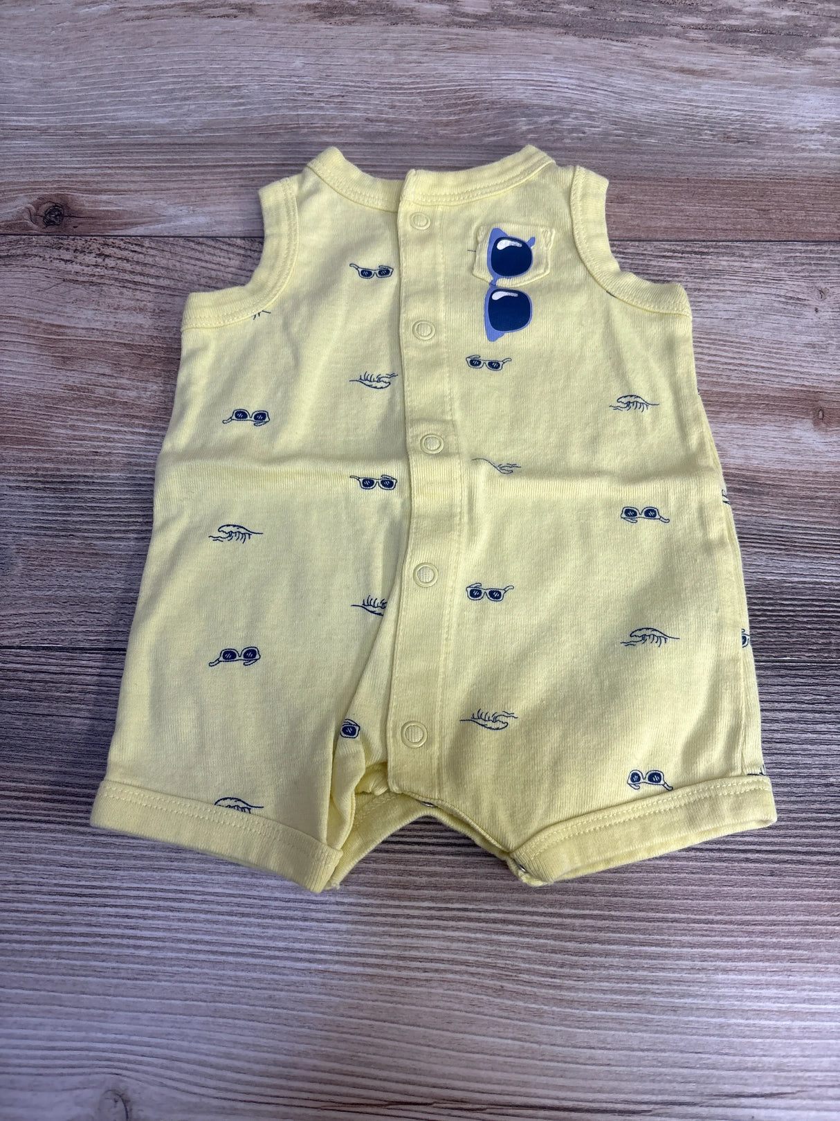 Top-down view of yellow newborn shortie romper with navy sunglass prints, front snap closure, sunglass patch on wood surface