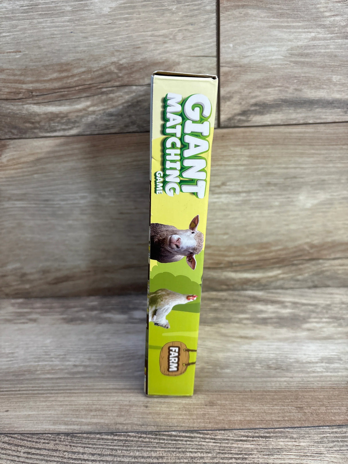 Side view of Giant Matching Card Game box with yellow spine, Giant Matching text, and sheep and chicken on wood surface