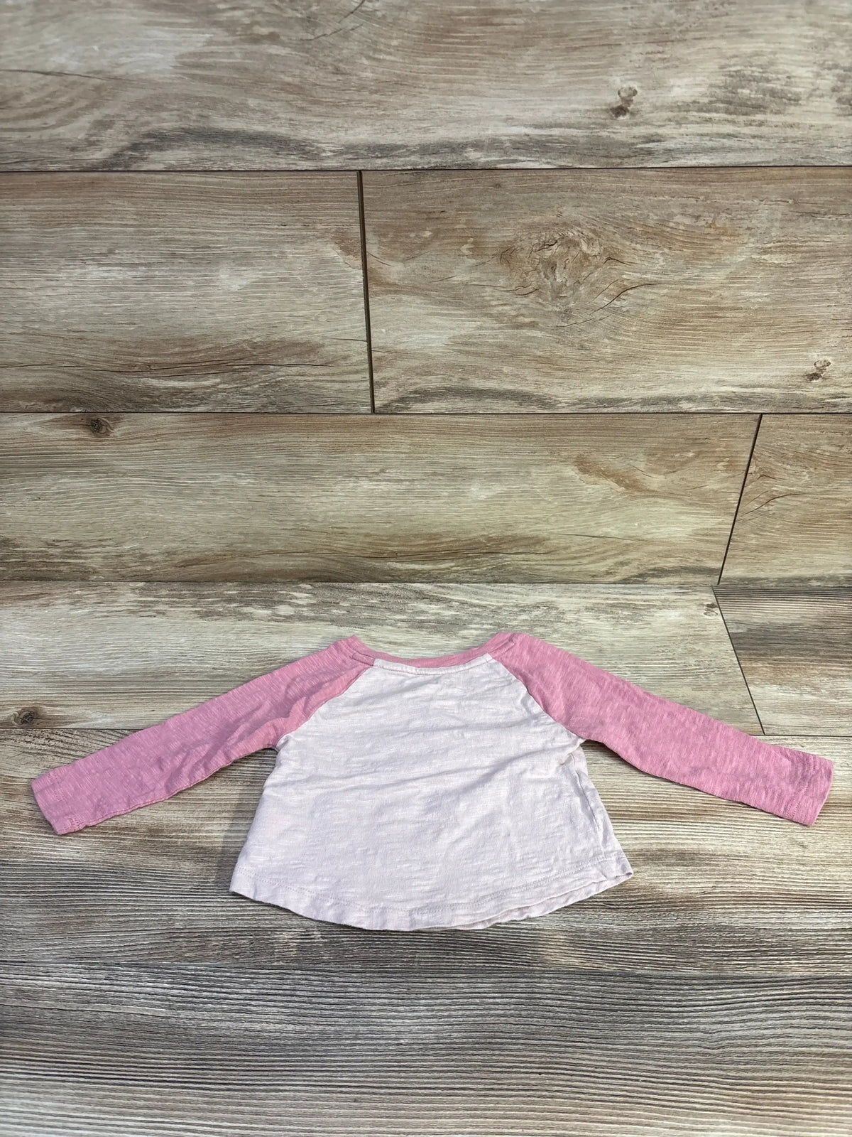 Top-down view of Baby Gap x Sesame Street raglan tee showcasing pink long sleeves, white body, and crew neckline on wood plank background
