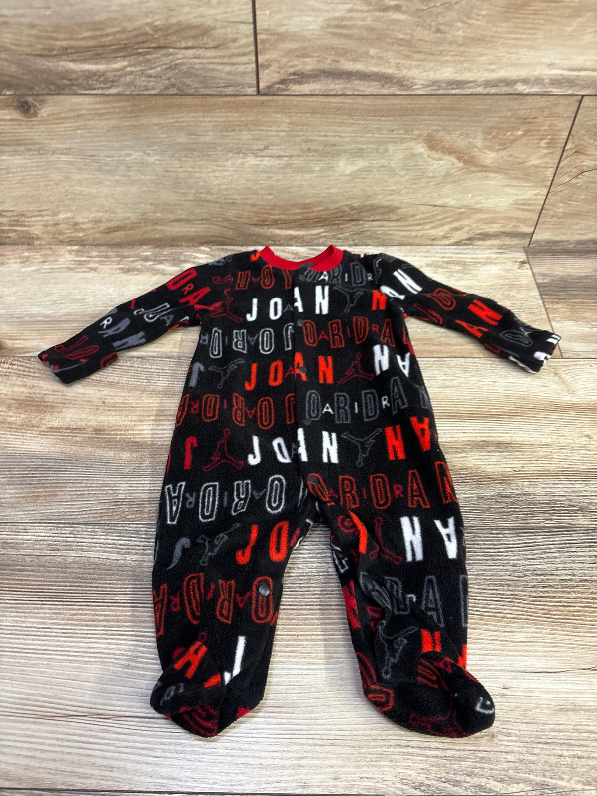 Front view of black Jordan blanket snap sleeper showcasing logo pattern, footed legs, and snap closures on a wood-look floor