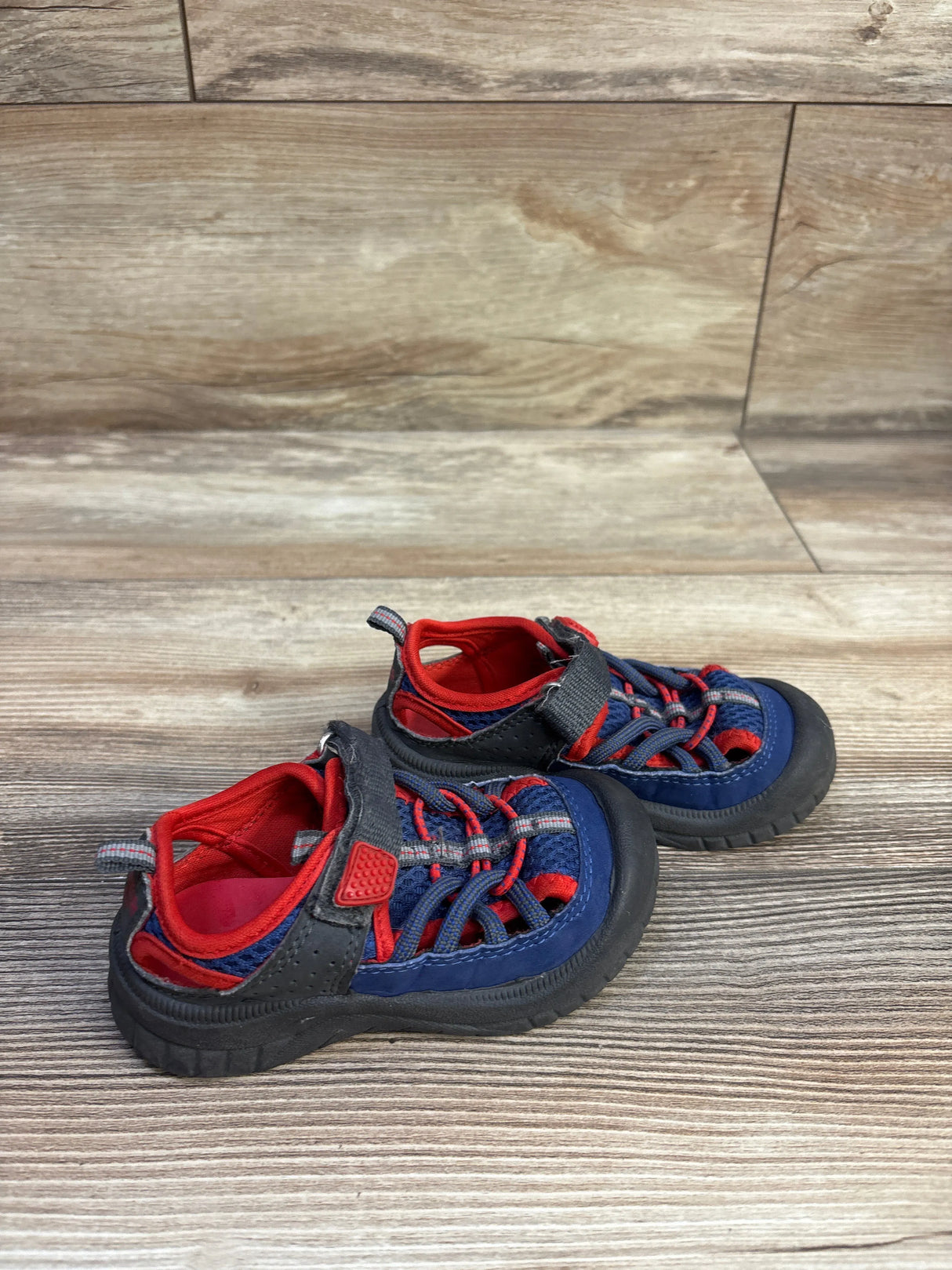 Angled view of Oshkosh toddler bump-toe sandal, navy and red, showing bungee laces, Velcro strap, and black toe bumper on wood