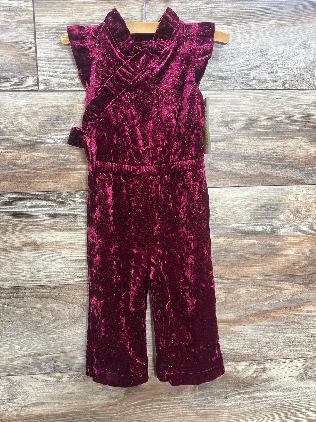 Front view of toddler girl jumpsuit in burgundy crushed velvet showcasing flutter sleeves wrap bodice and wide leg on wooden hanger against wood plank floor