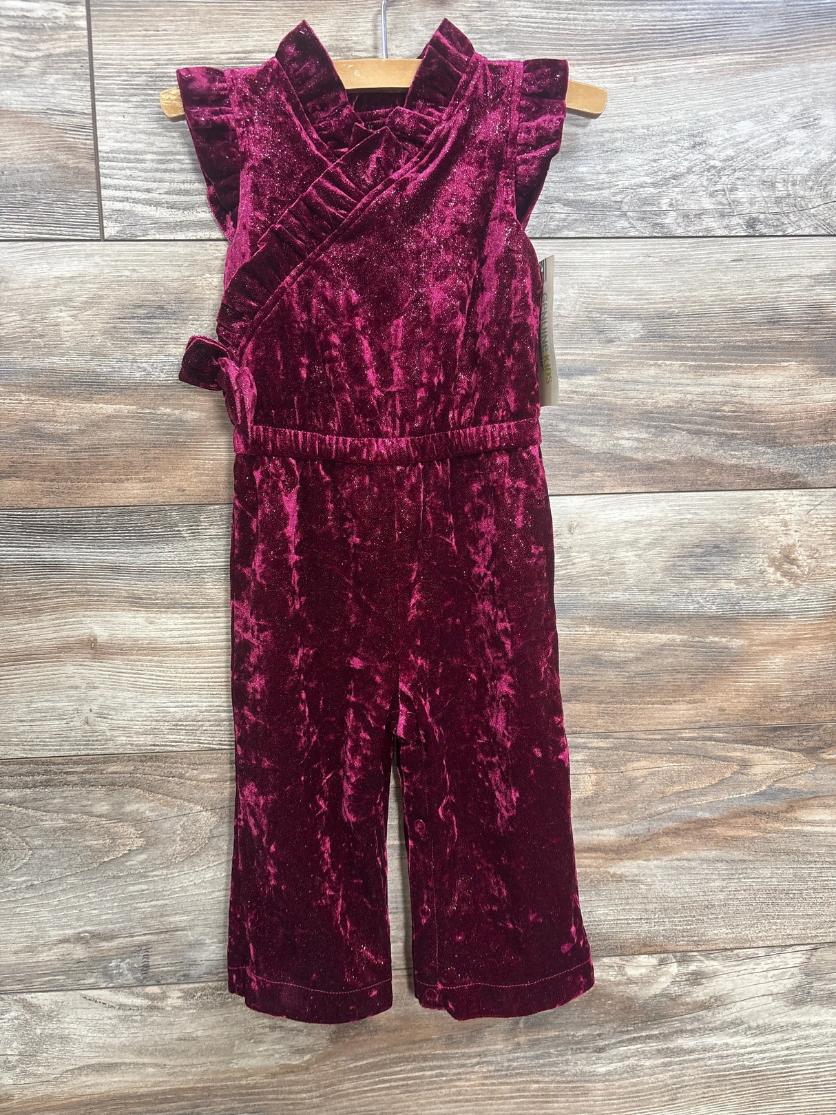Front view of toddler girl jumpsuit in burgundy crushed velvet showcasing flutter sleeves wrap bodice and wide leg on wooden hanger against wood plank floor