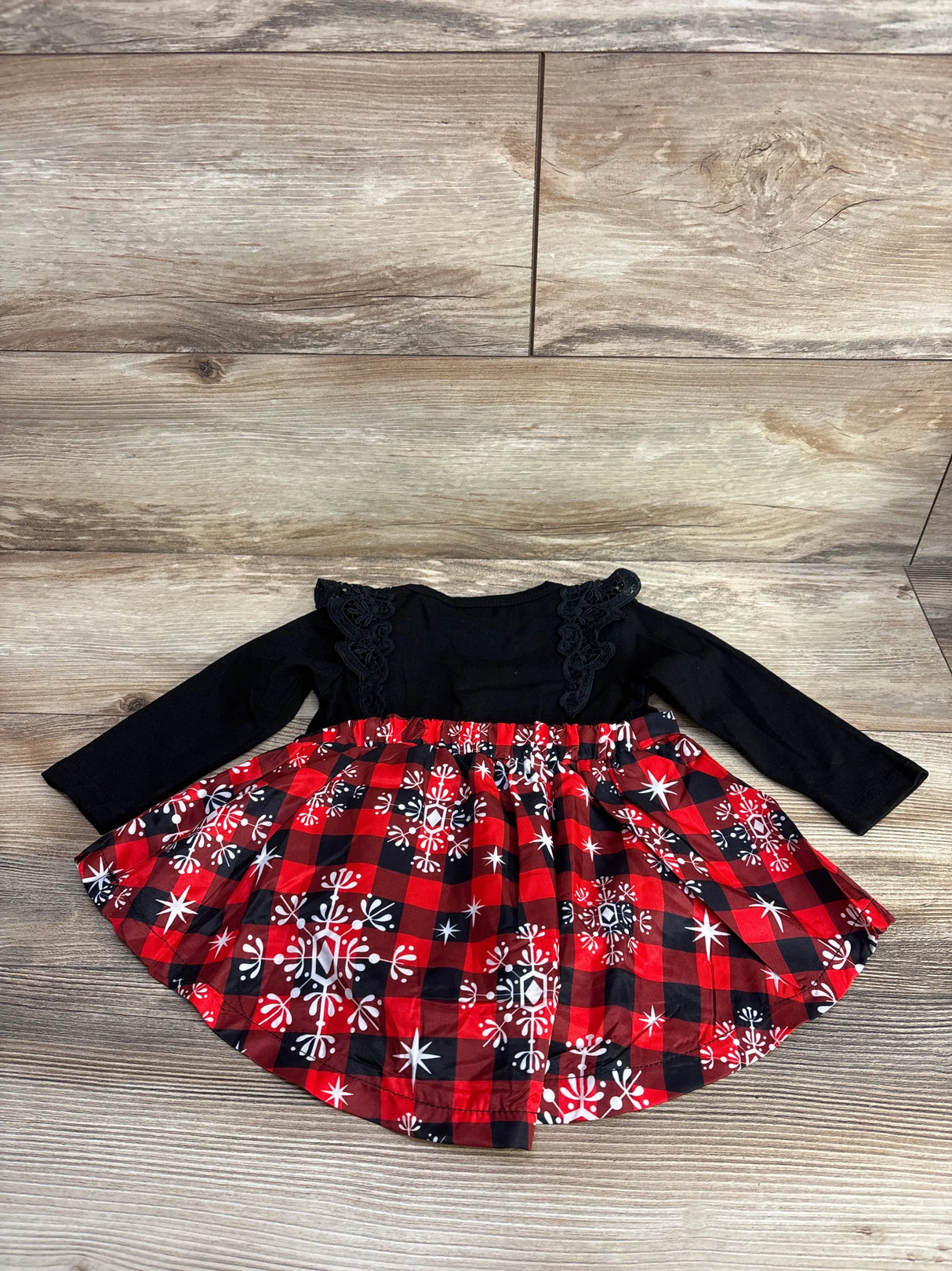 Baby long sleeve dress with black top and red plaid snowflake skirt, laid on wood floor