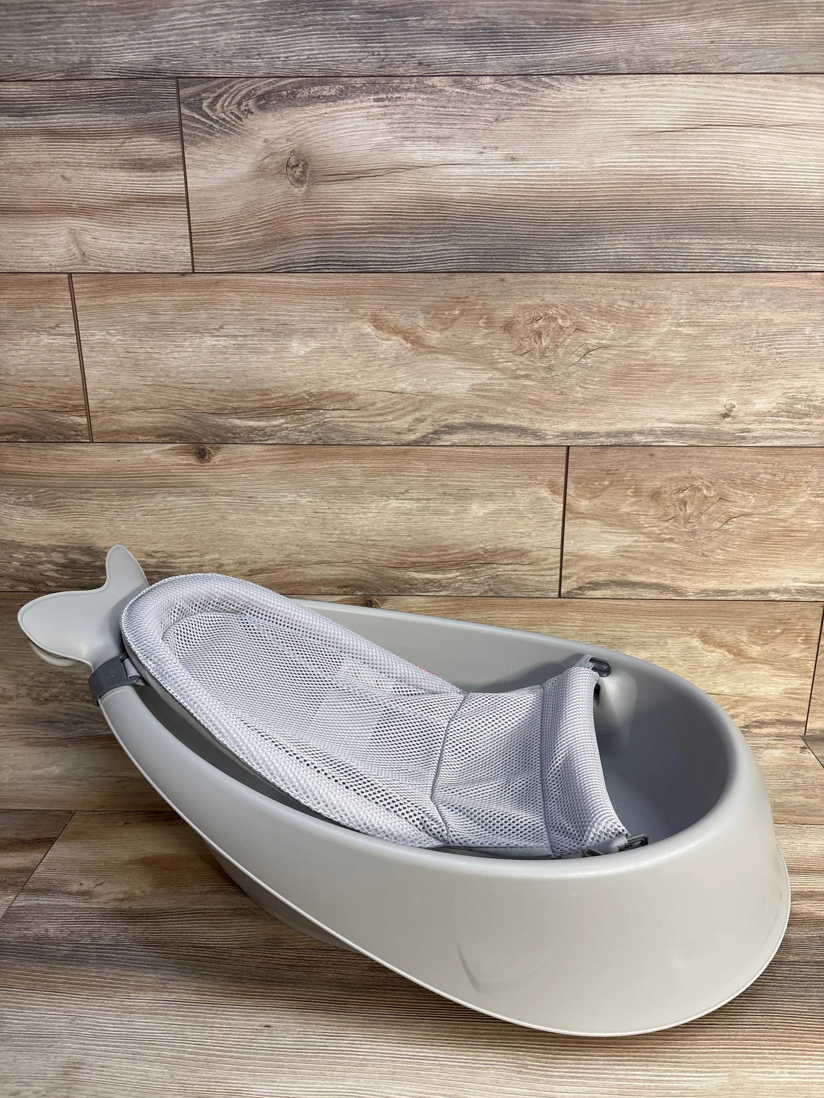Angled view of Skip Hop Moby Smart Sling 3 in 1 baby bathtub in gray showcasing its mesh Smart Sling on a wood floor