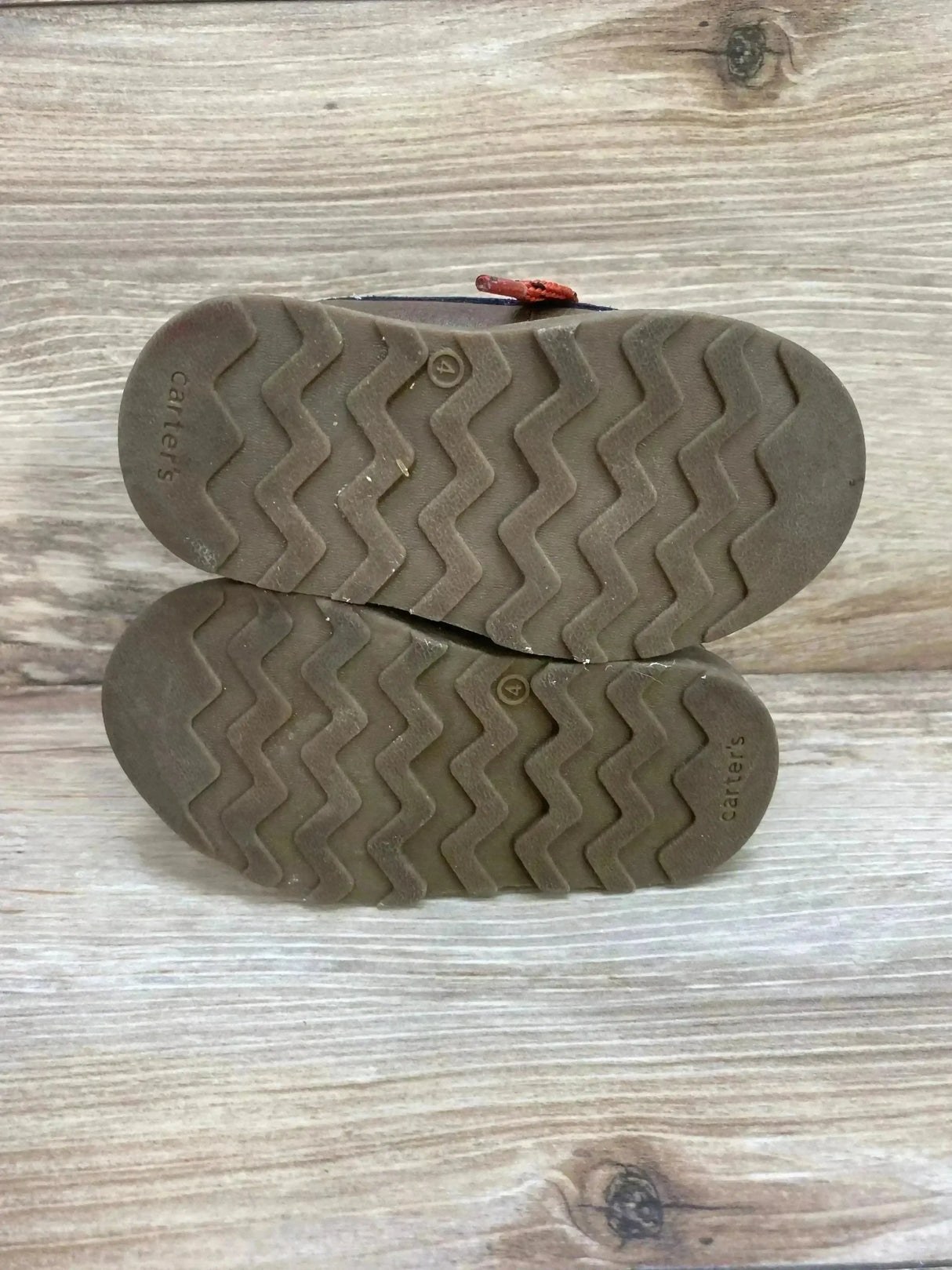 Bottom view of Carter’s Norman toddler ankle boots showing brown rubber soles with zigzag tread on wood background