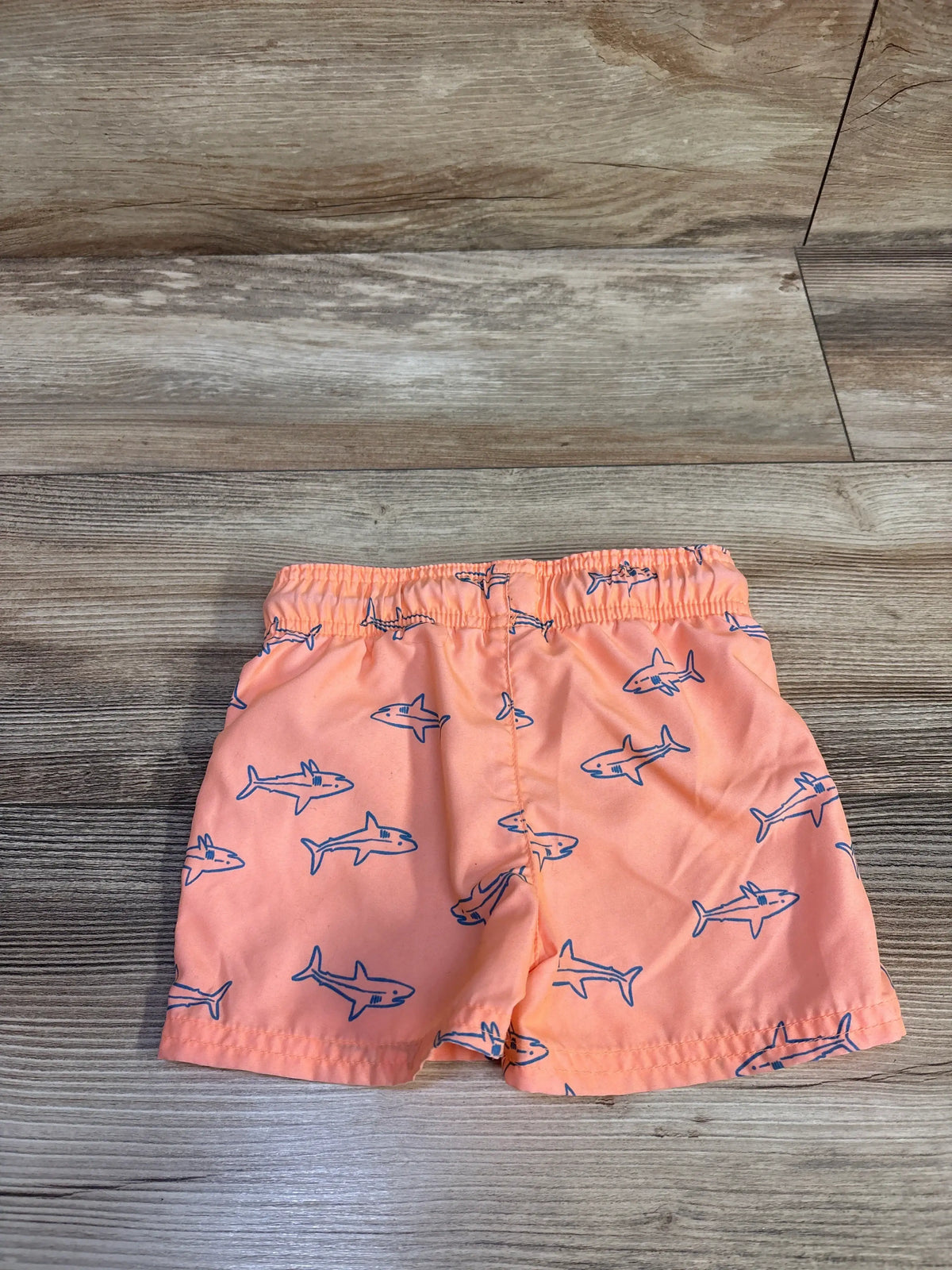 Front view of Cat and Jack orange shark swim trunks in size 18 months on a wood floor