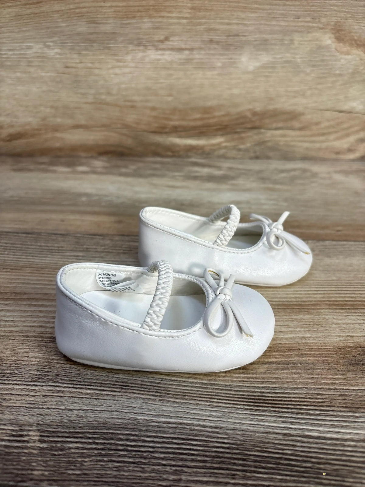Front view of white Janie and Jack soft sole ballet flats showcasing bow details and elastic straps on a wood grain surface