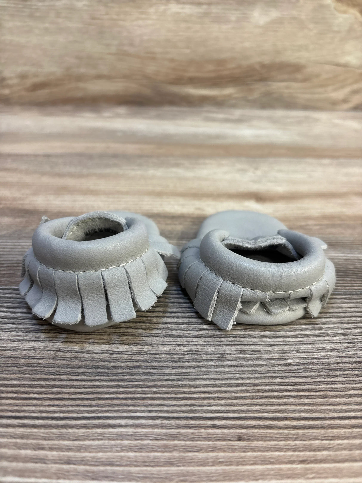 Close-up of Freshly Picked Classic Mocc Grey sz 0c showcasing pair of gray leather baby moccasins with fringe trim on wood plank floor