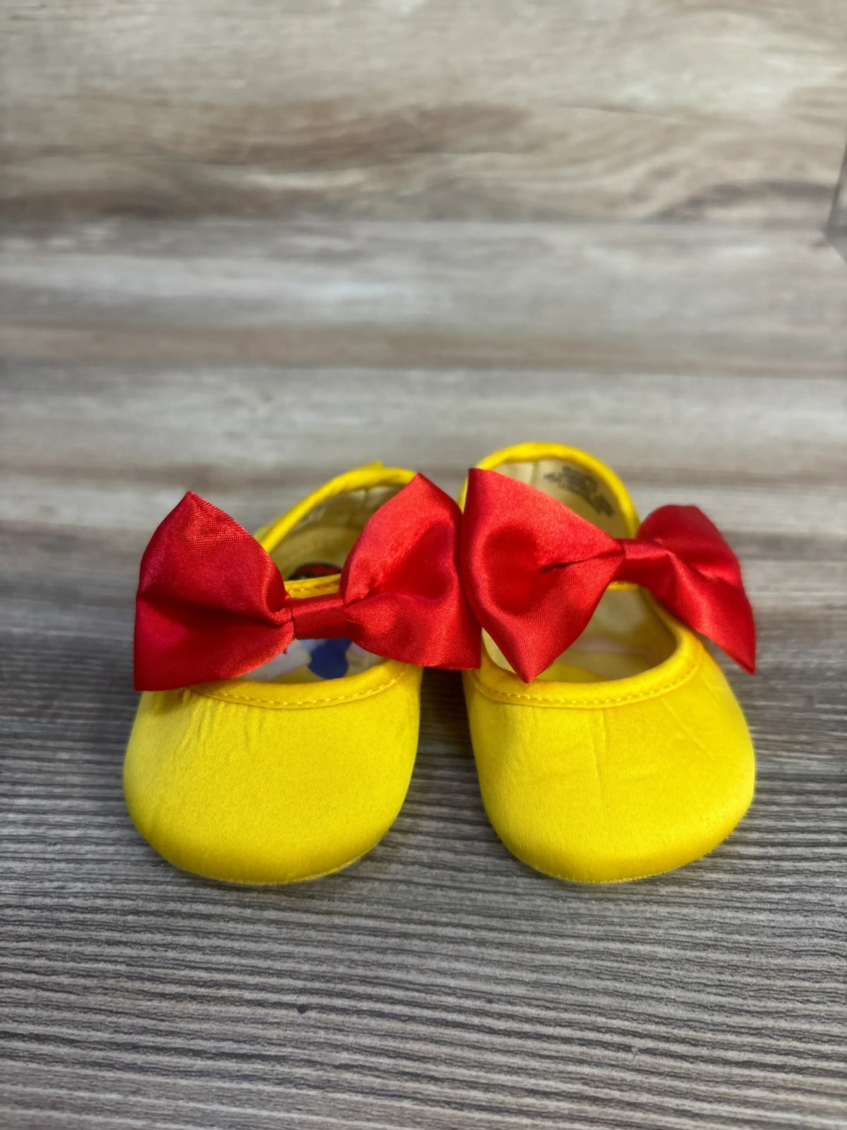 Front view of Disney Baby Snow White soft sole shoes in yellow showcasing red bows with a wood-grain background