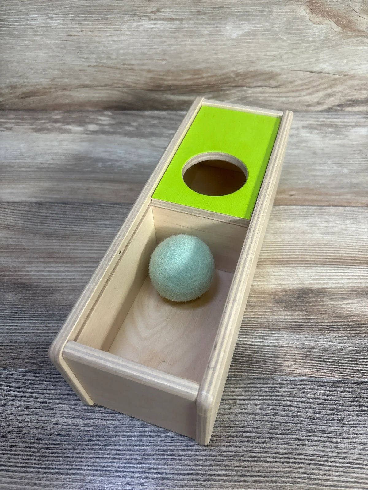 Angled view of Lovevery Sliding Top Box showcasing its green sliding top, circular opening, and felt ball on a wood-grain surface