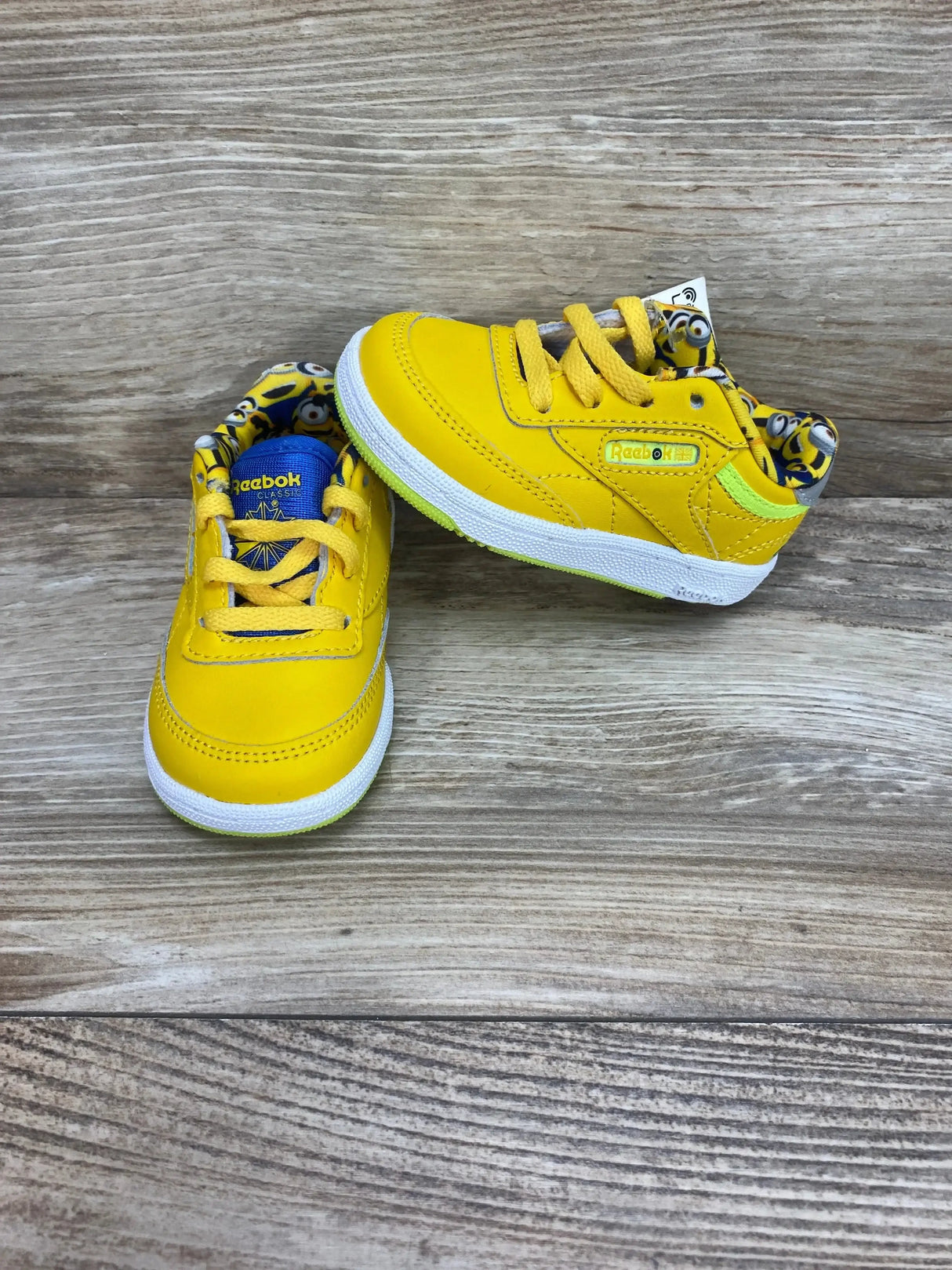 Three-quarter view of NEW Illuminations Minions x Reebok Club C 85 Toddler Sneakers in yellow with white sole and Minions tongue patch on wood plank floor