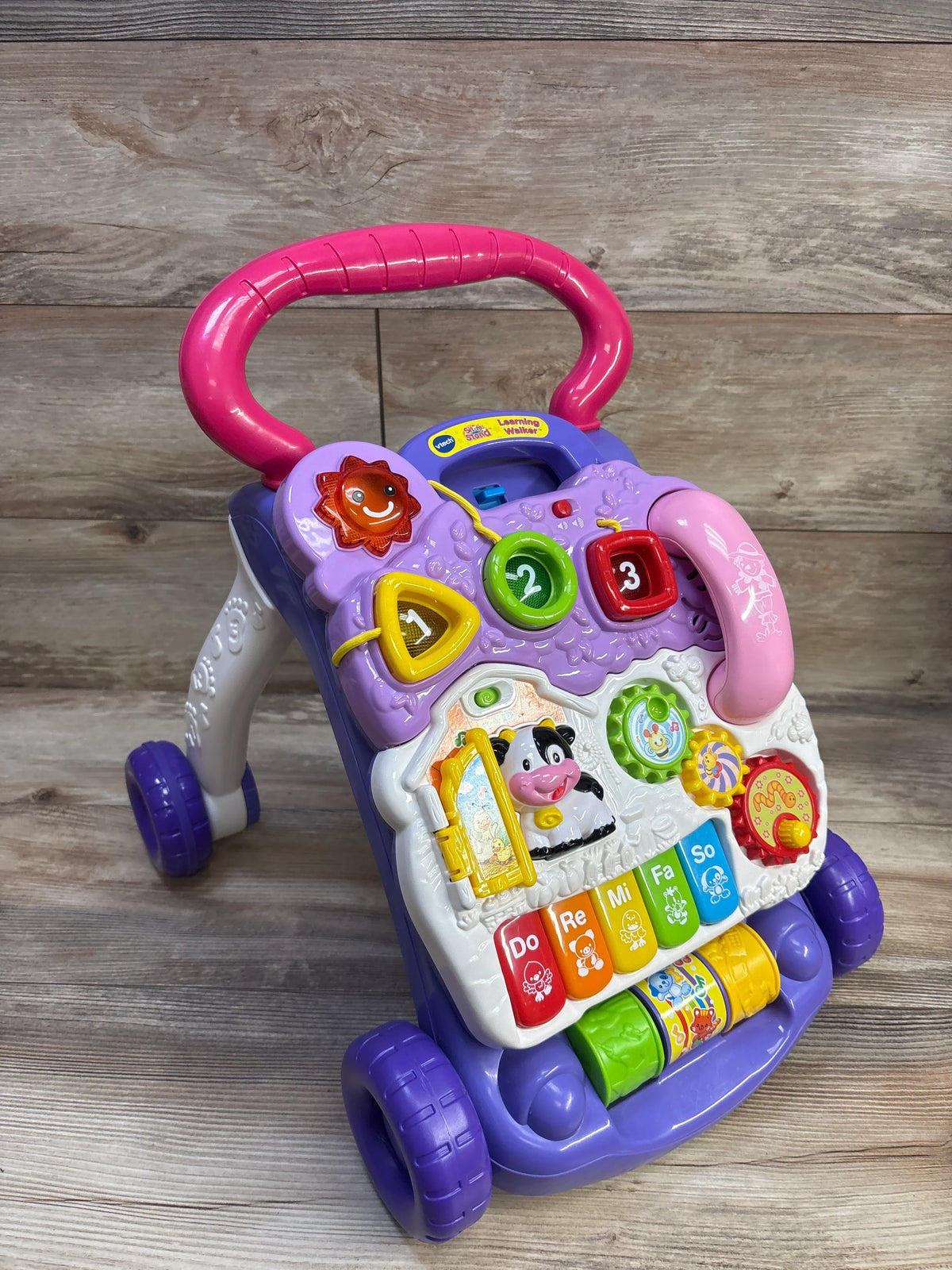 Three-quarter front view of VTech Sit-to-Stand Learning Walker showcasing colorful piano keys, shape sorters, spinning rollers, and purple-pink handle on wooden floor background