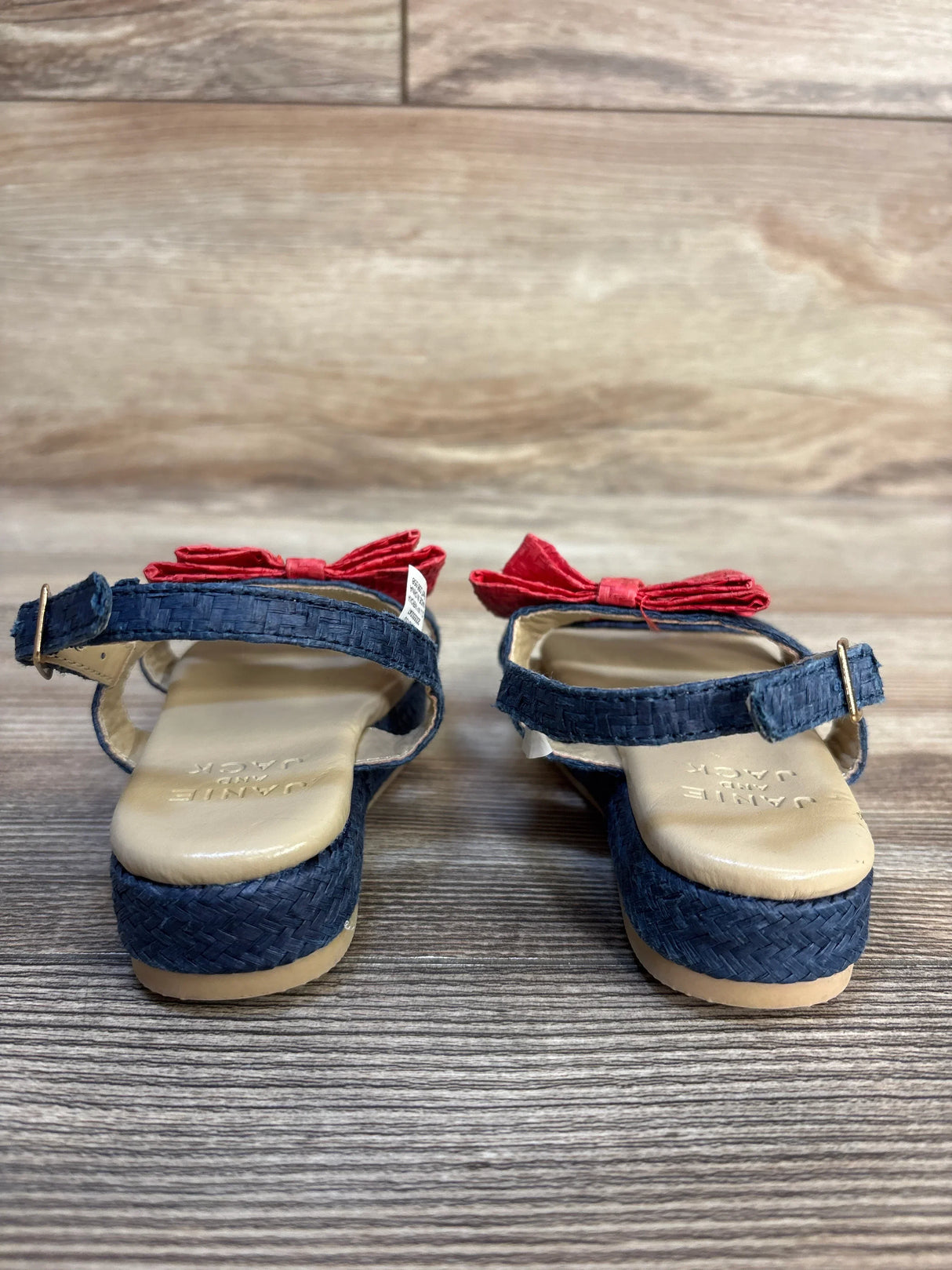 Pair of Janie & Jack Double Bow Straw Sandals in Merchant Marine showing red bows and ankle straps on wood floor