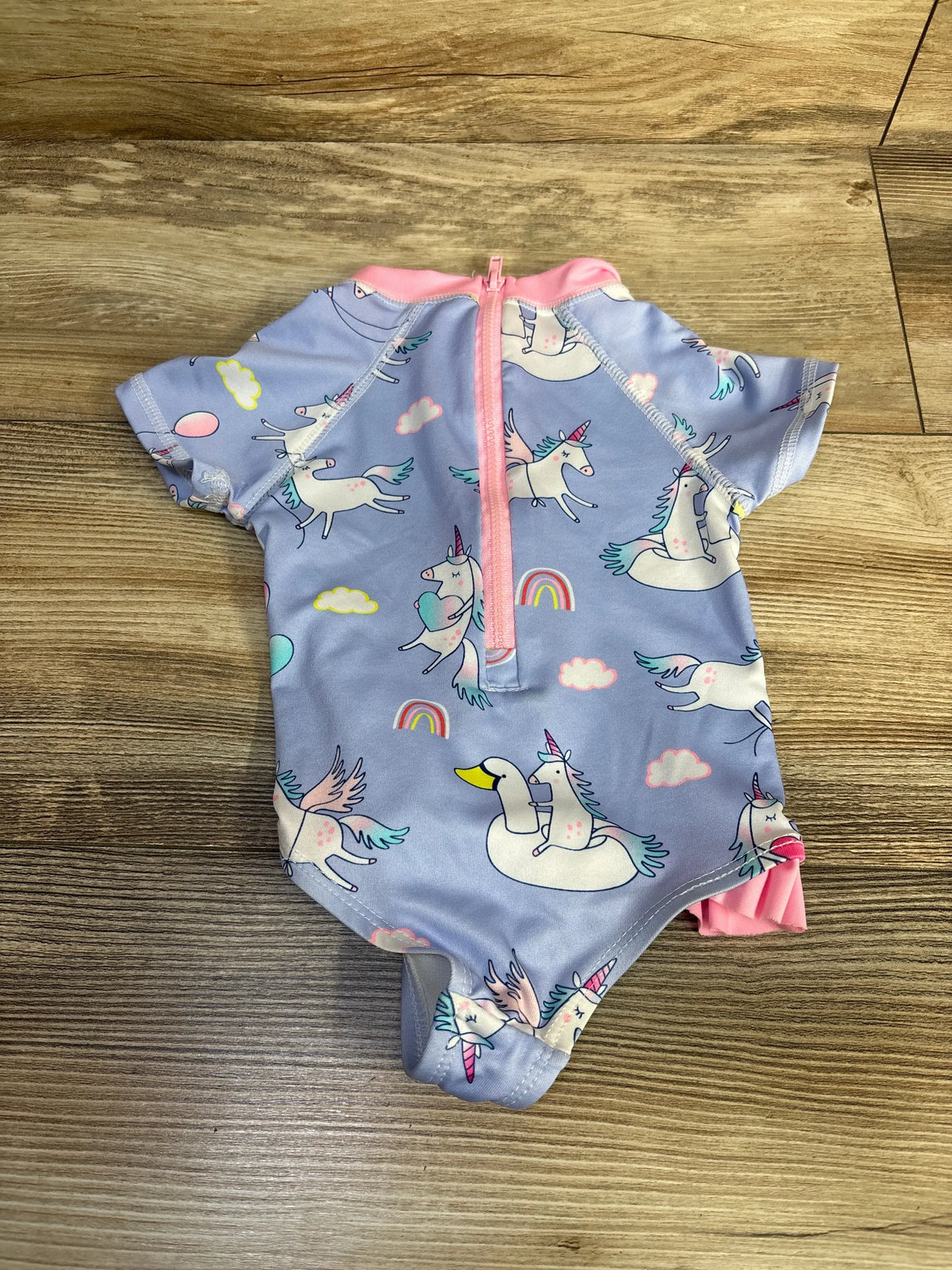 Flat view of Wippette one-piece unicorn rash guard swimsuit showcasing purple unicorn print and pink zipper on wood floor