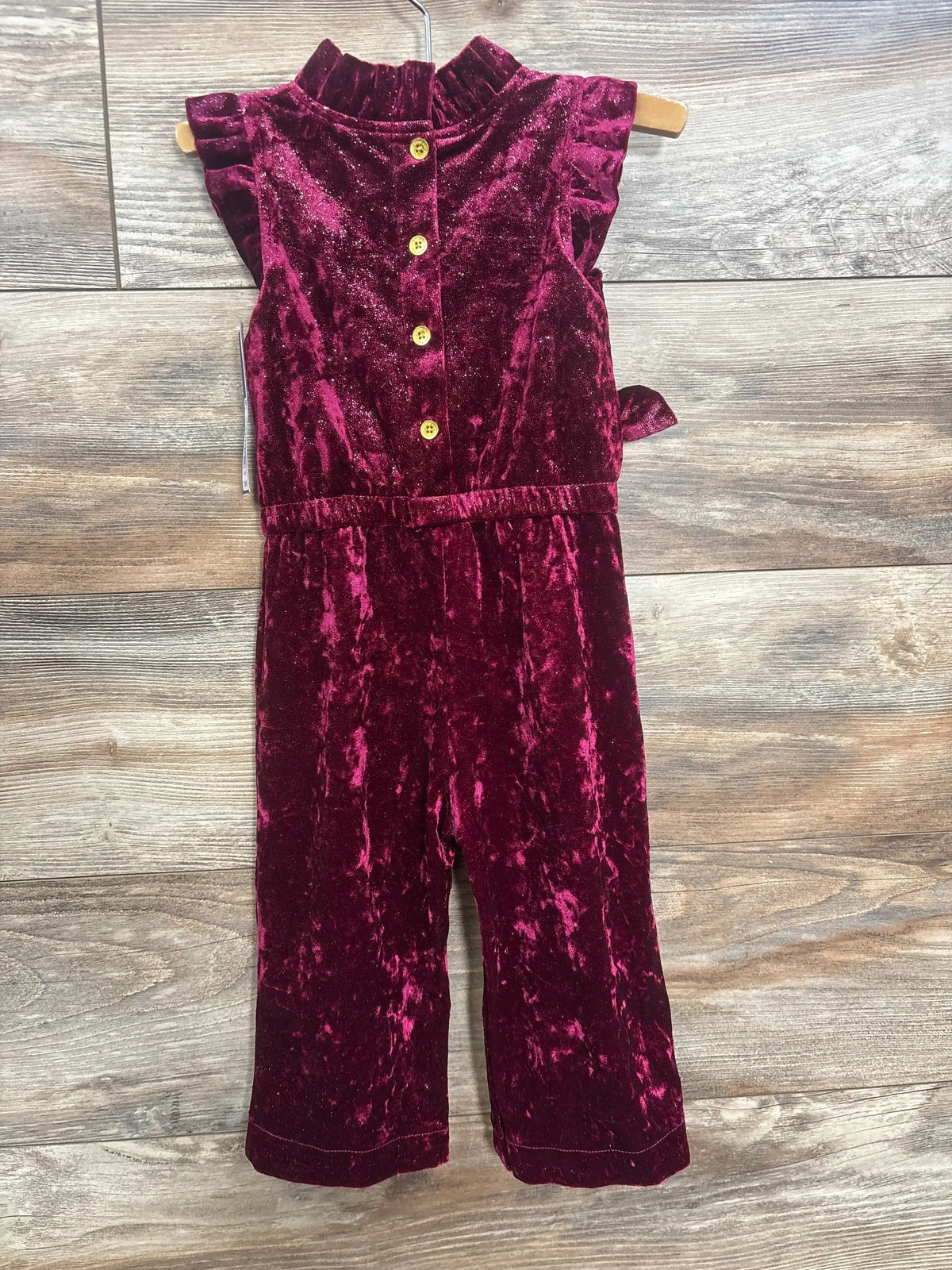 Flat lay of OshKosh velvet sparkle jumpsuit in burgundy with ruffle shoulders button front and tie waist on wood plank floor