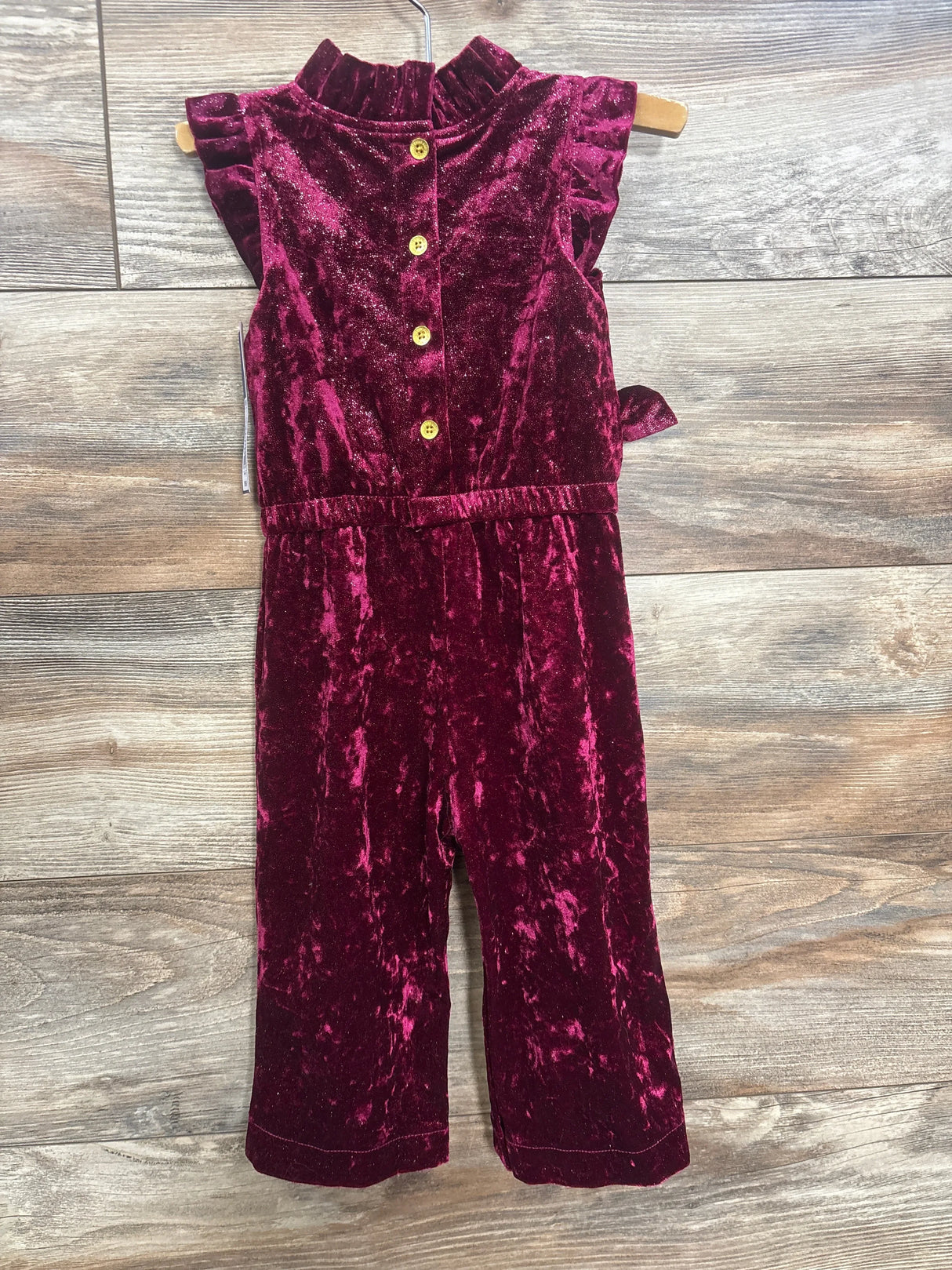 Flat lay of OshKosh velvet sparkle jumpsuit in burgundy with ruffle shoulders button front and tie waist on wood plank floor