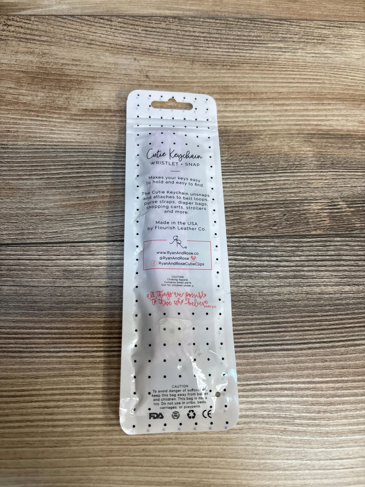Top-down view of Cutie Keychain wristlet in retail packaging showing black polka-dot design and product information on a wooden surface
