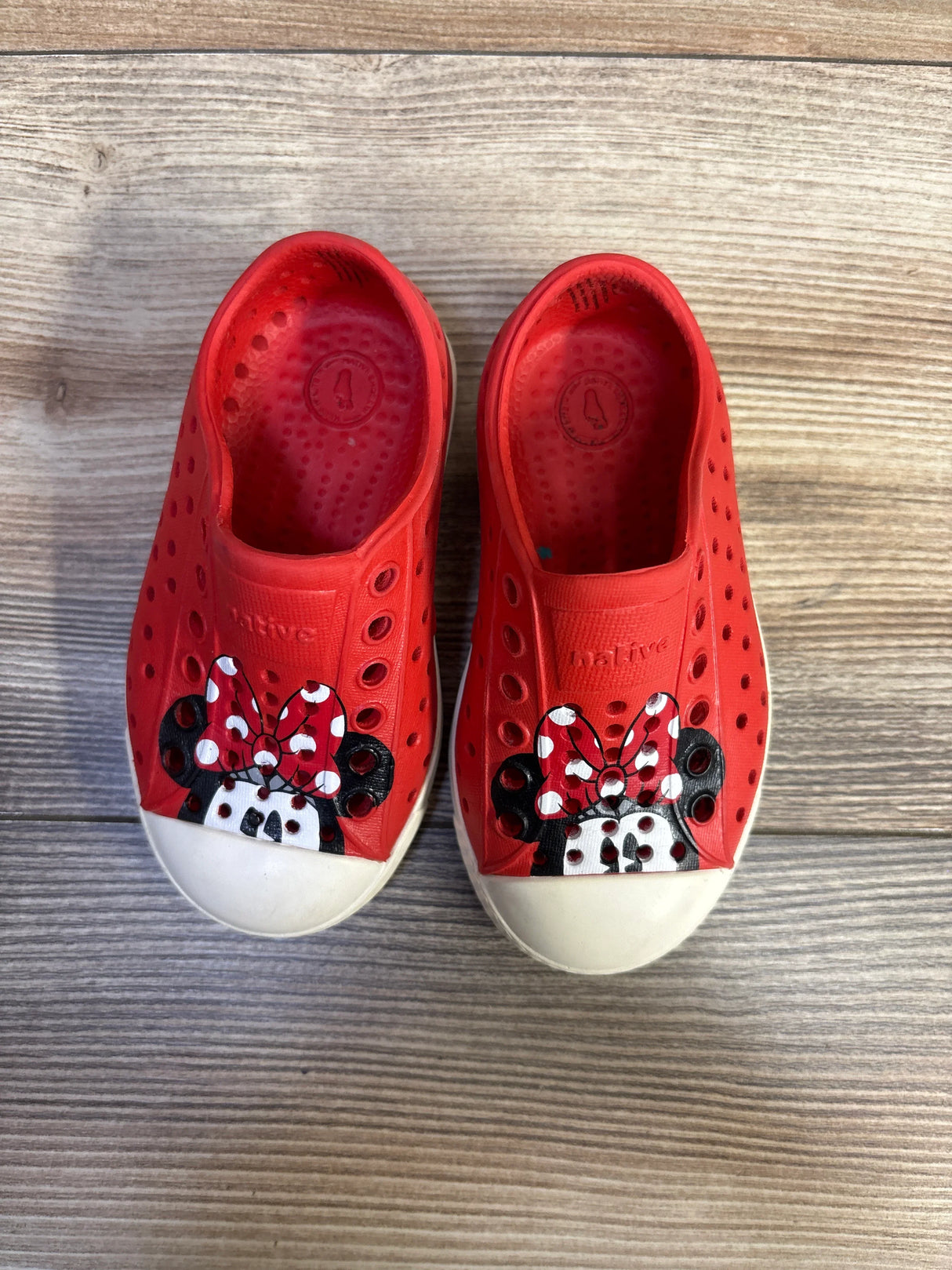 Top-down view of red Native Jefferson slip-on toddler shoes showing painted Minnie Mouse faces on the toes with white toe caps on a wood floor