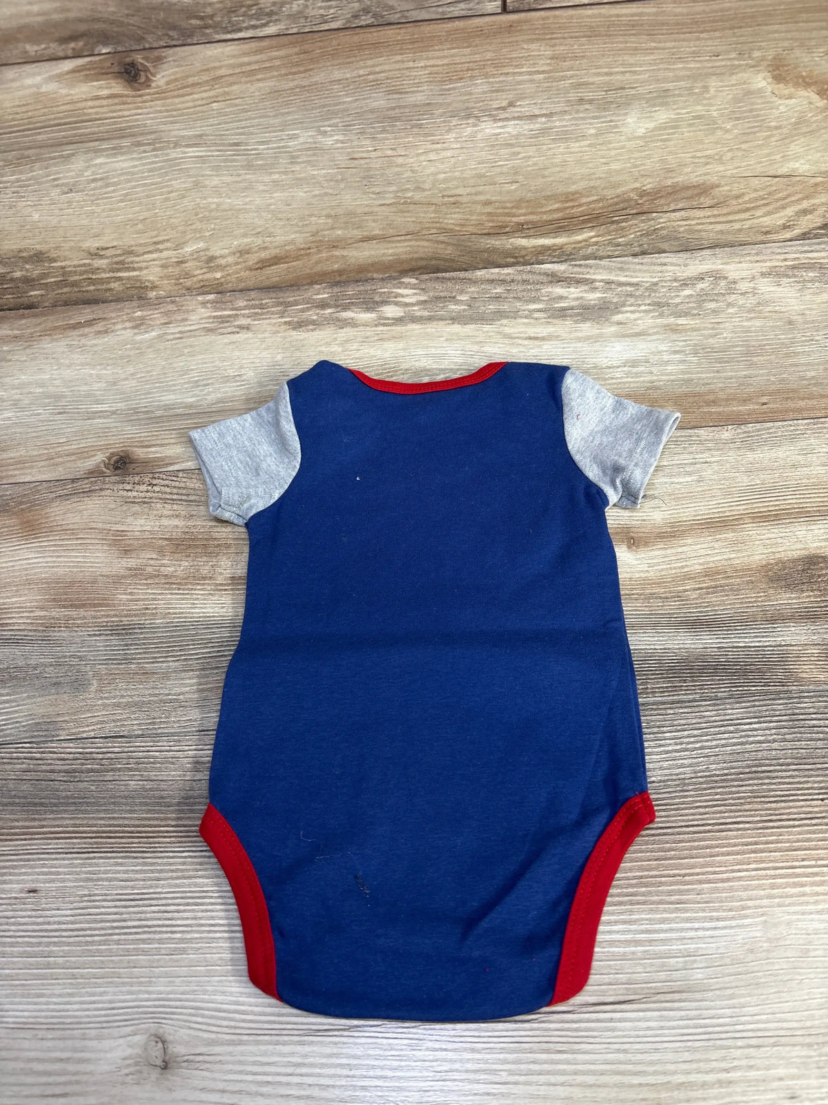 Front view of blue Guess baby bodysuit showcasing gray short sleeves and red trim on a wood background
