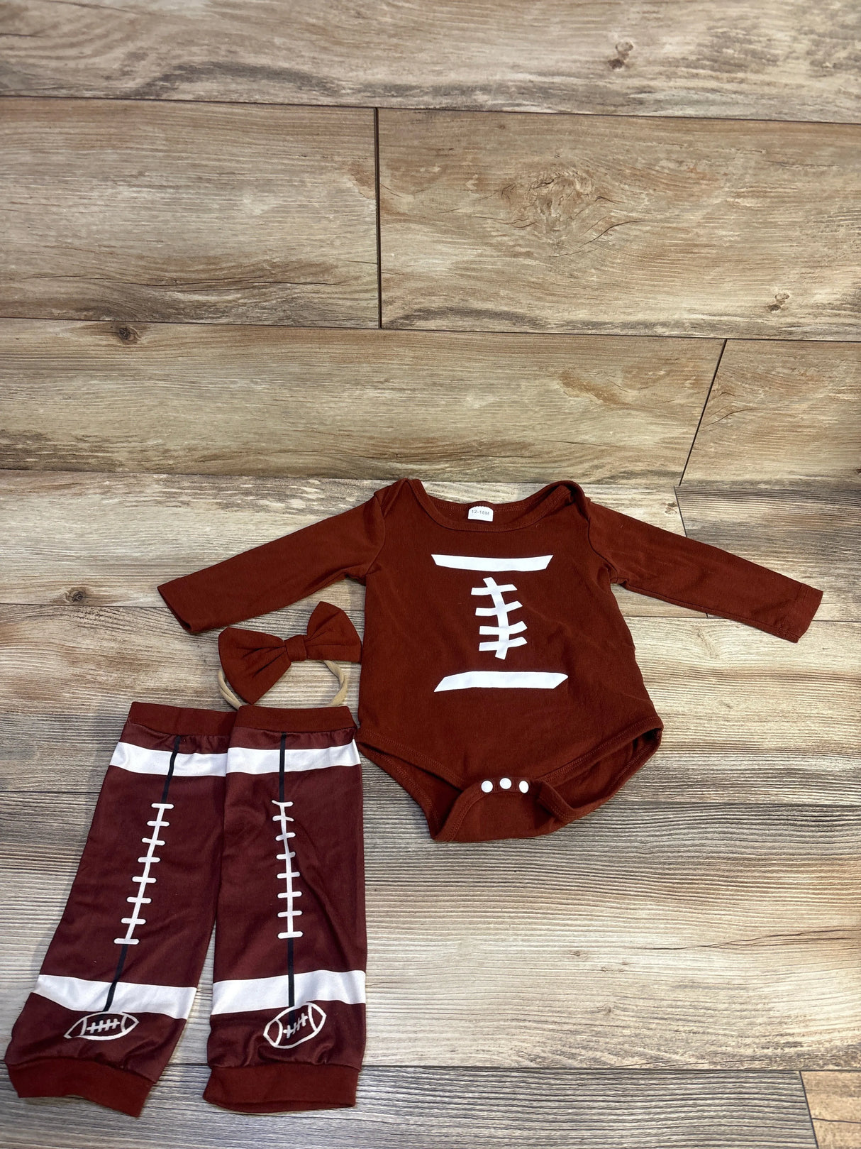 Flat lay of brown football-themed bodysuit set with white laces print, matching leg warmers and bow headband on wood background