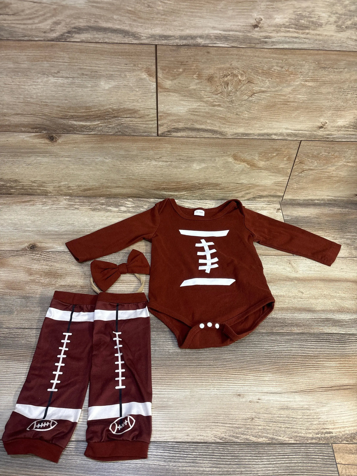 Flat lay of brown football-themed bodysuit set with white laces print, matching leg warmers and bow headband on wood background