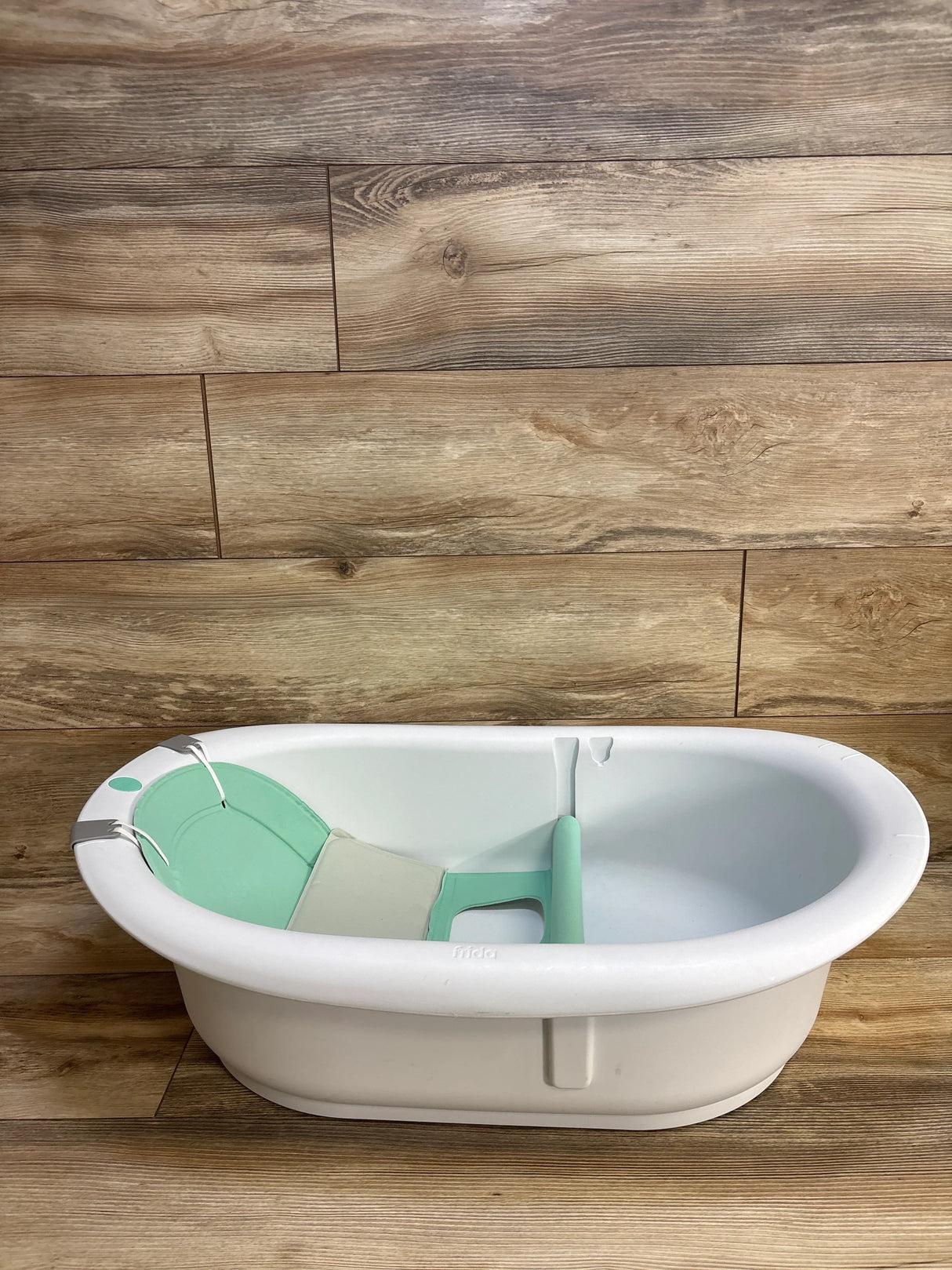 Side view of Frida Baby 4-in-1 Grow-With-Me tub with mint-green infant support insert and divider on wood floor