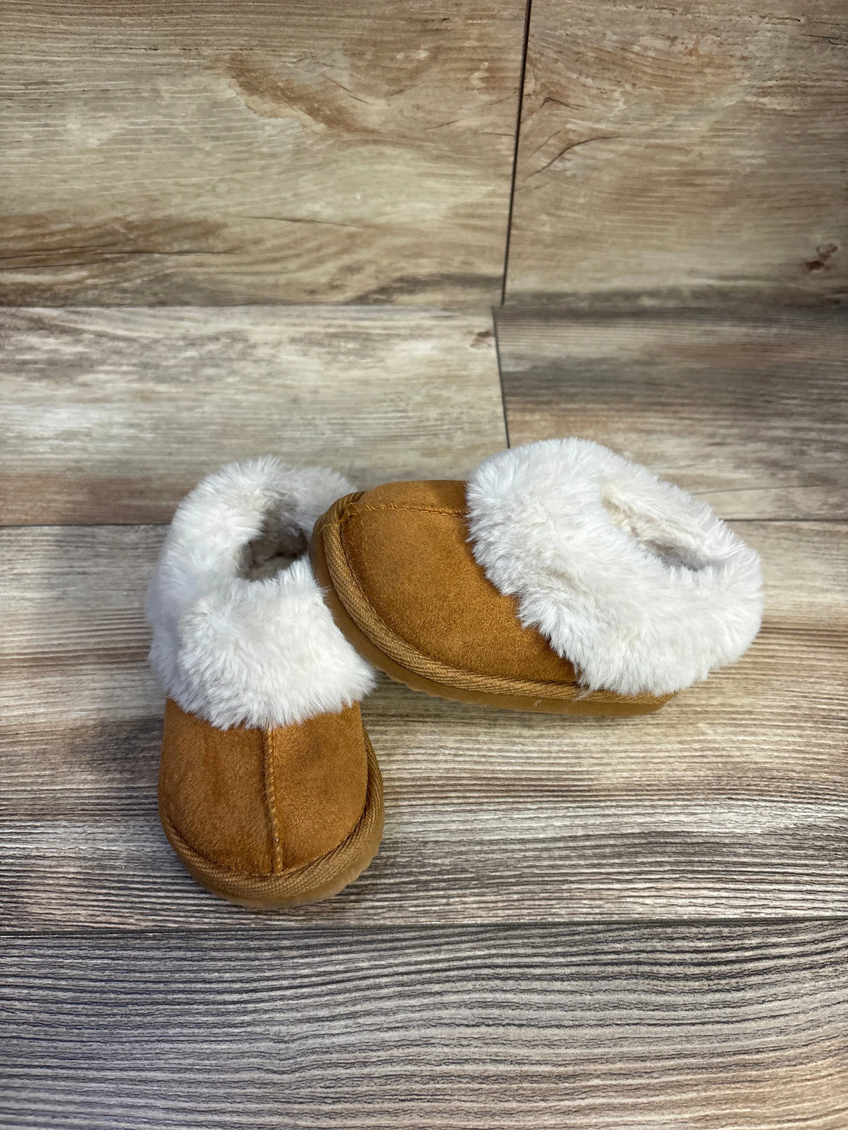 Angled top view of brown toddler slippers with white faux-fur cuff, stitched toe, plush sole on light wood background