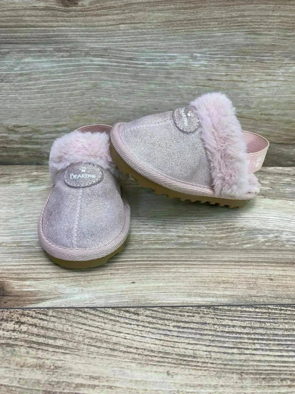 Angled front view of BEARPAW Rebecca light pink suede slippers with sheepskin cuff, elastic slingback, and ridged sole on wood