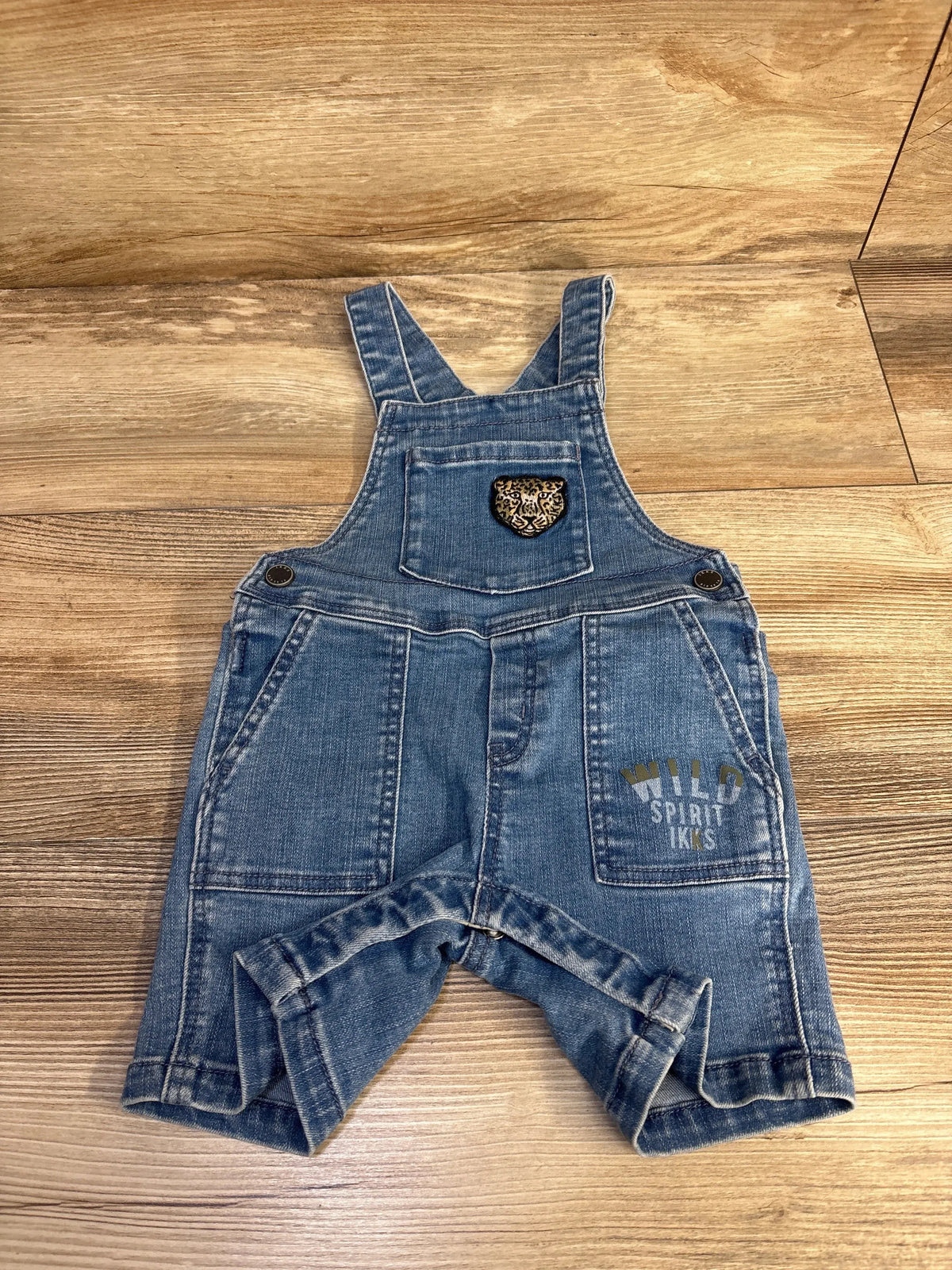 Flat lay of blue toddler denim shortalls 18 months showcasing front bib pocket with leopard patch, WILD SPIRIT IKKS print, and side buttons on wood background