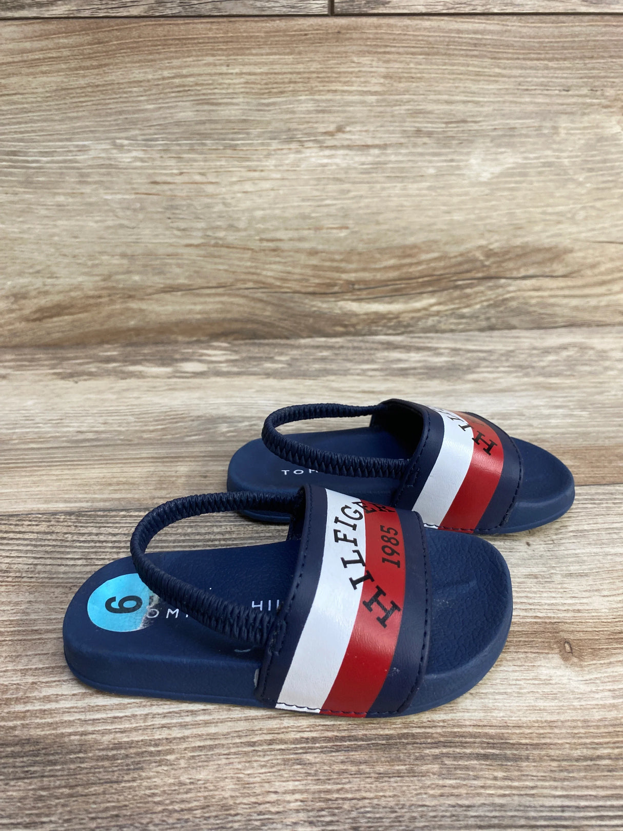 Navy blue toddler sandals with red and white Tommy Hilfiger stripes on wood background