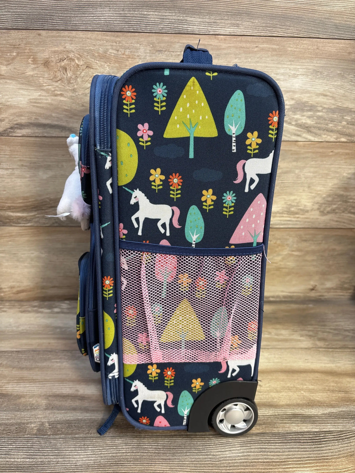 Front three-quarter view of Crckt Kids Softside Carry On Suitcase showcasing unicorn print and front mesh pocket on a wood floor